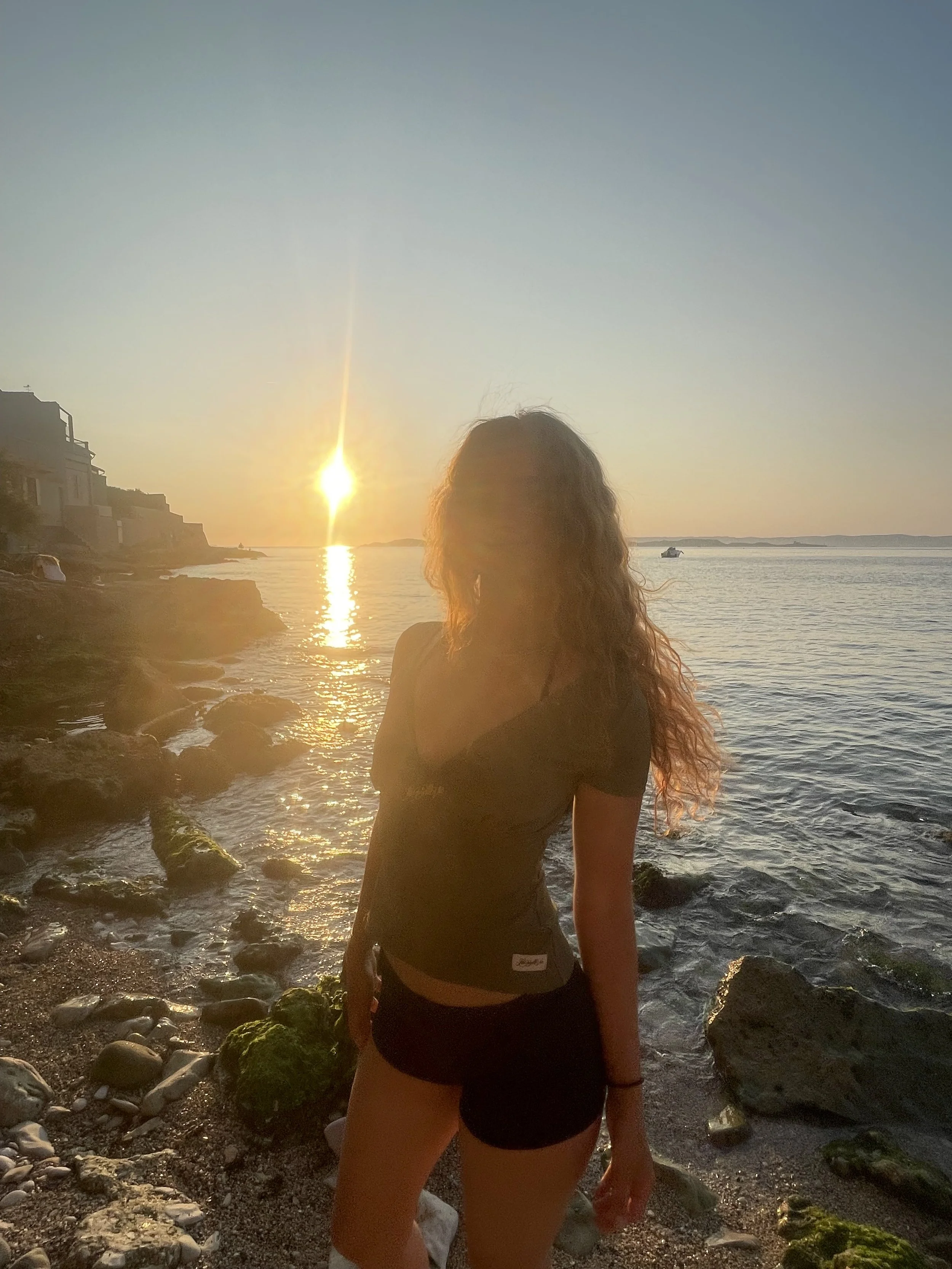 Une femme aux cheveux longs et bouclés se tient sur une plage rocheuse au coucher du soleil, avec la mer en arrière-plan, des bâtiments en bord de mer à gauche et un bateau à droite.
