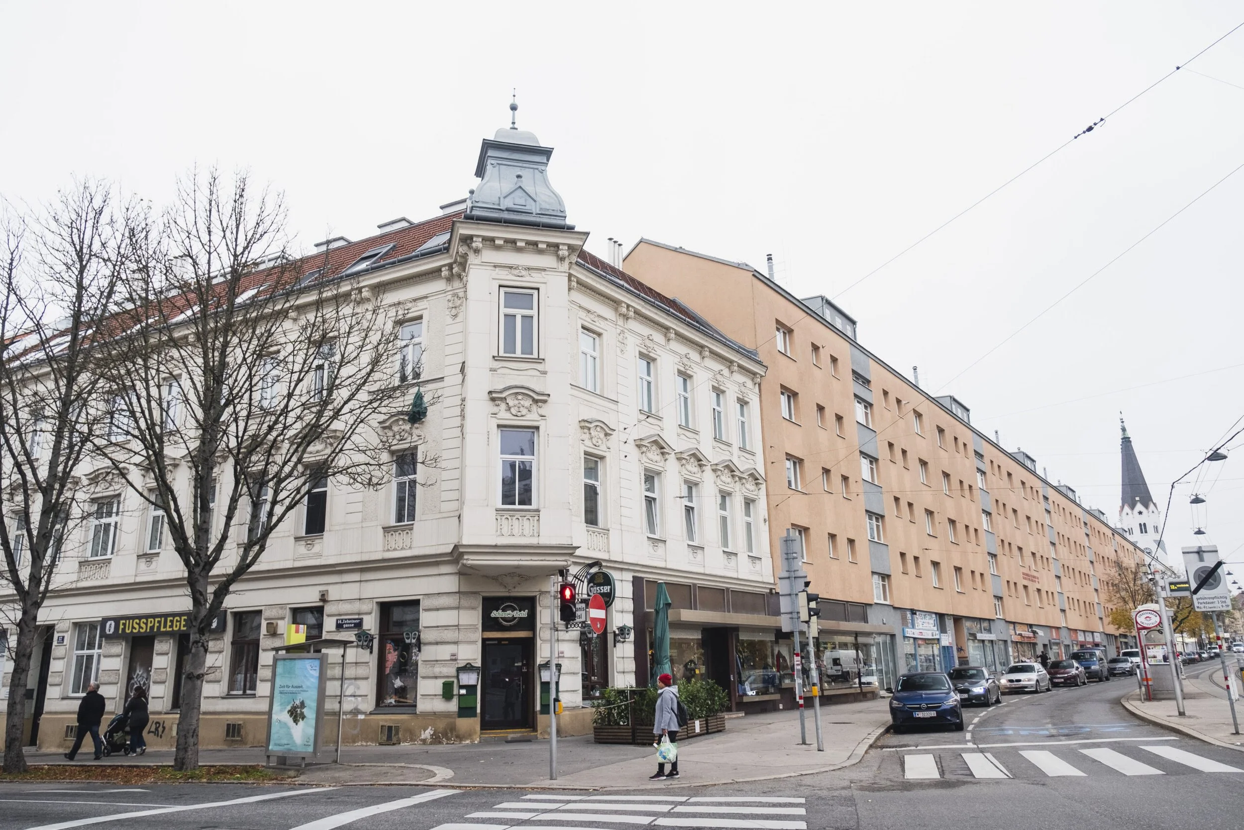 Stadtstraße mit historischen Gebäuden, Geschäften und Menschen an einem Gehweg. Im Hintergrund ist eine Kirche zu sehen.