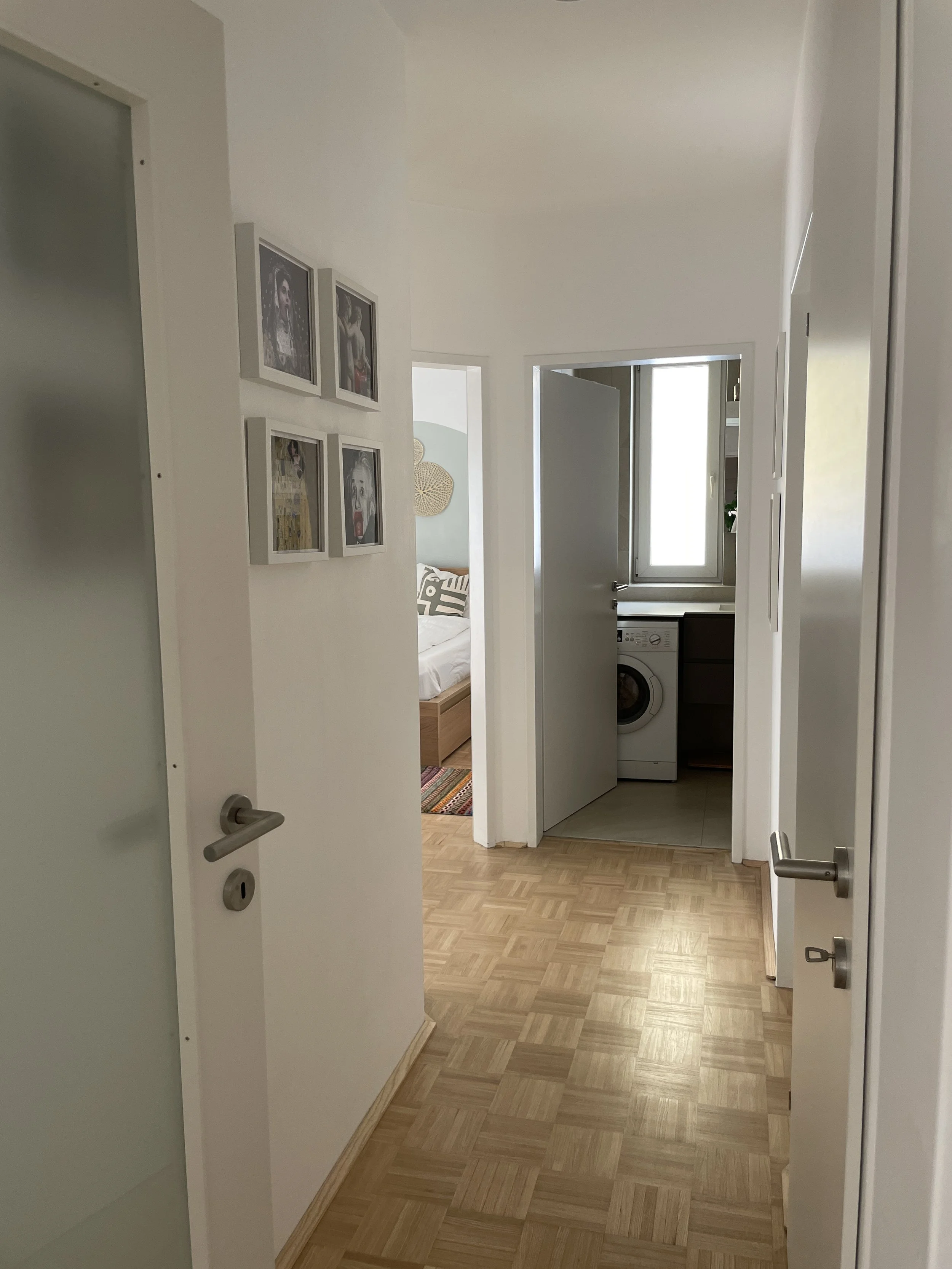 Flur mit Holzboden, offenen Türen, gerahmten Bildern an der Wand, Waschmaschine im Hintergrund und Schlafzimmer links.