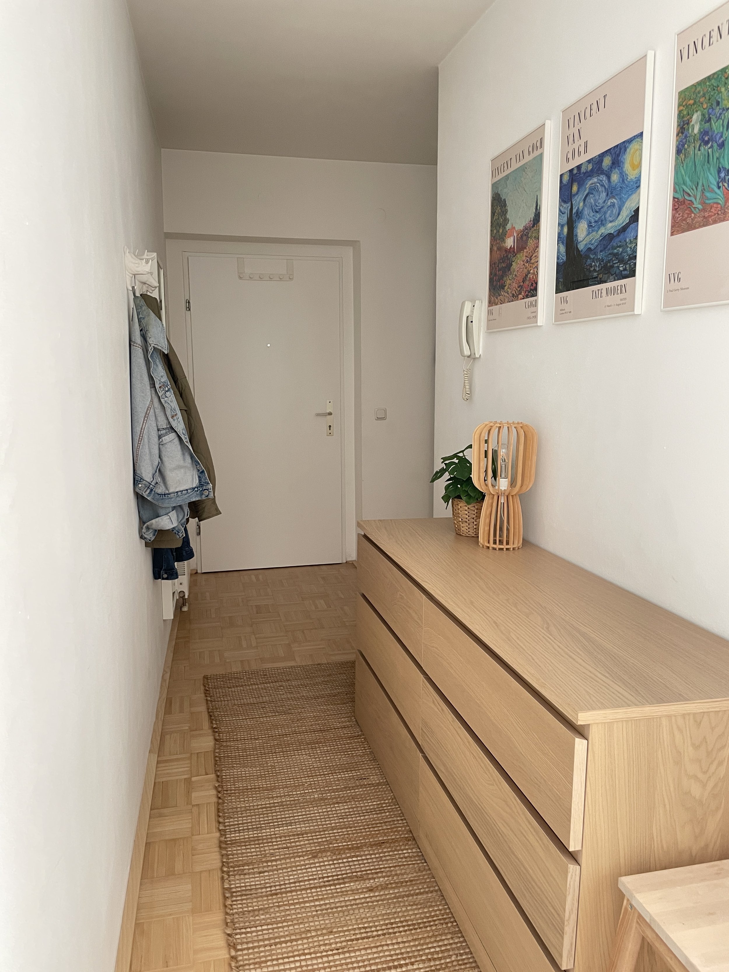 Flur mit weißer Wand, Holzboden, Holzkommode, Kleiderhaken mit Kleidung, beige Fußmatte, und Kunstposter an der Wand.