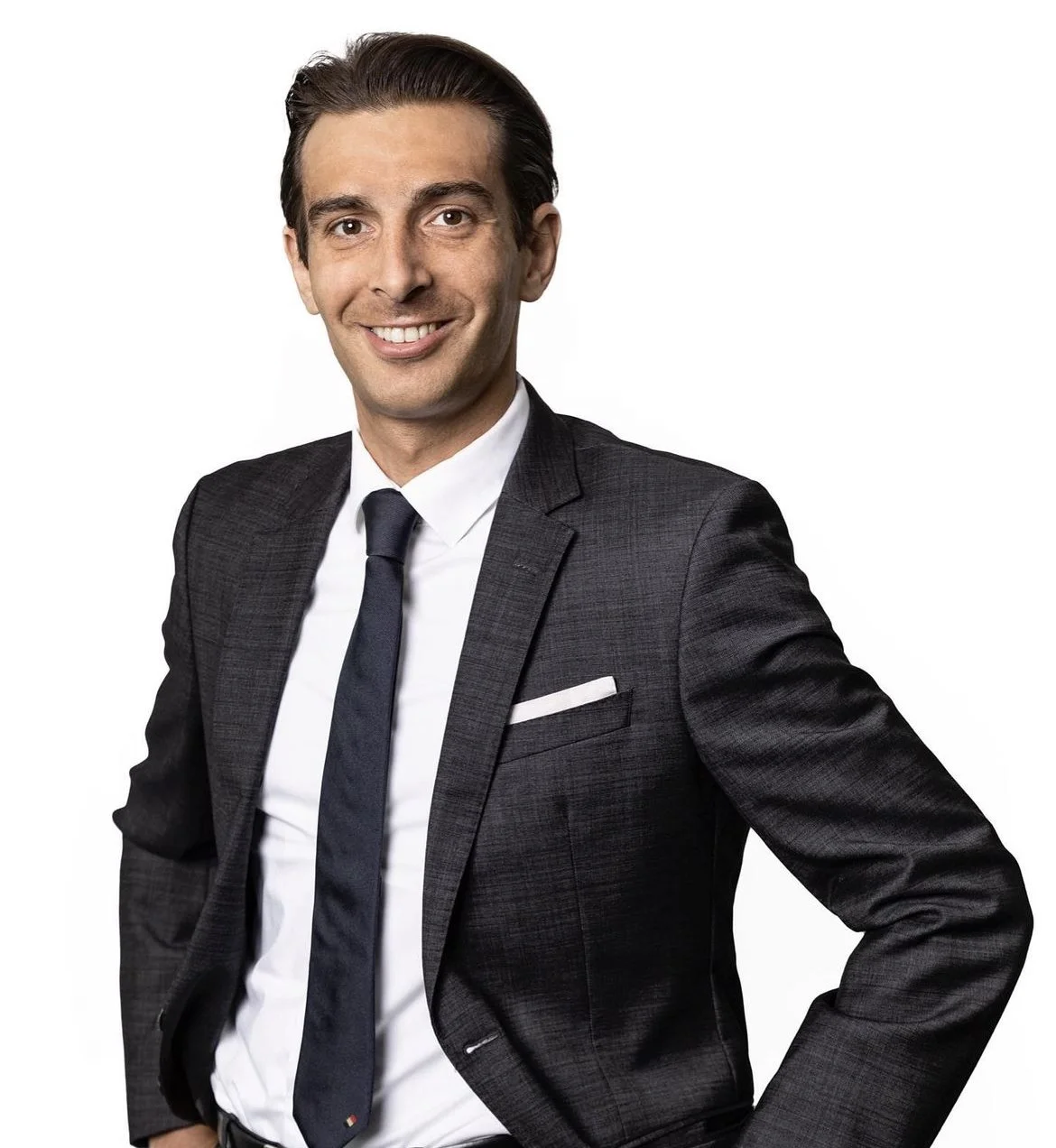 Ein Mann in einem dunklen Anzug mit weißem Hemd und Krawatte lächelt vor einem weißen Hintergrund.