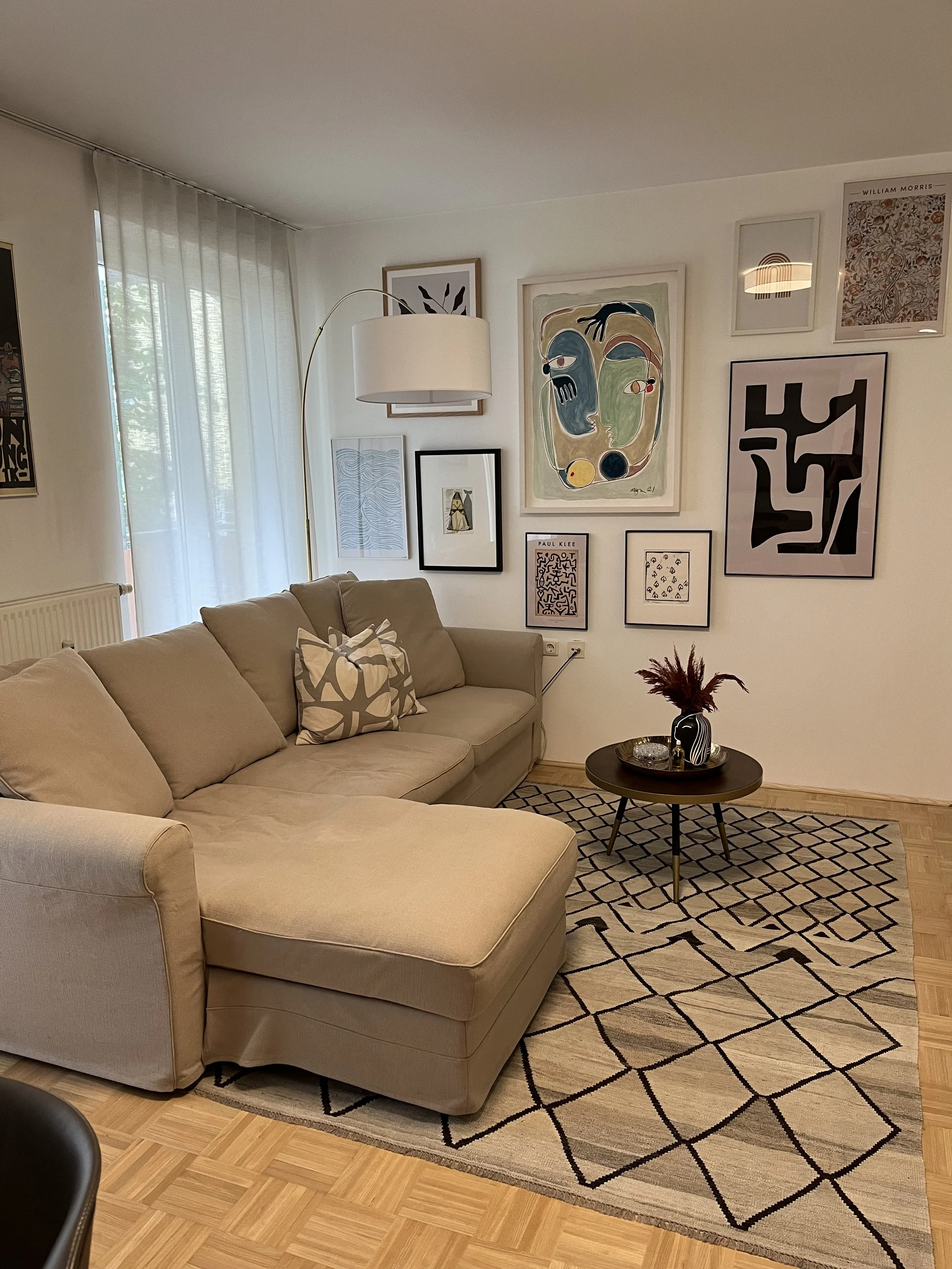Modernes Wohnzimmer mit beigem Sofa, dekoriertem Kissen, Stehlampe und abstrakten Kunstwerken an der Wand. Ein Couchtisch mit Dekorationen steht auf einem gemusterten Teppich.