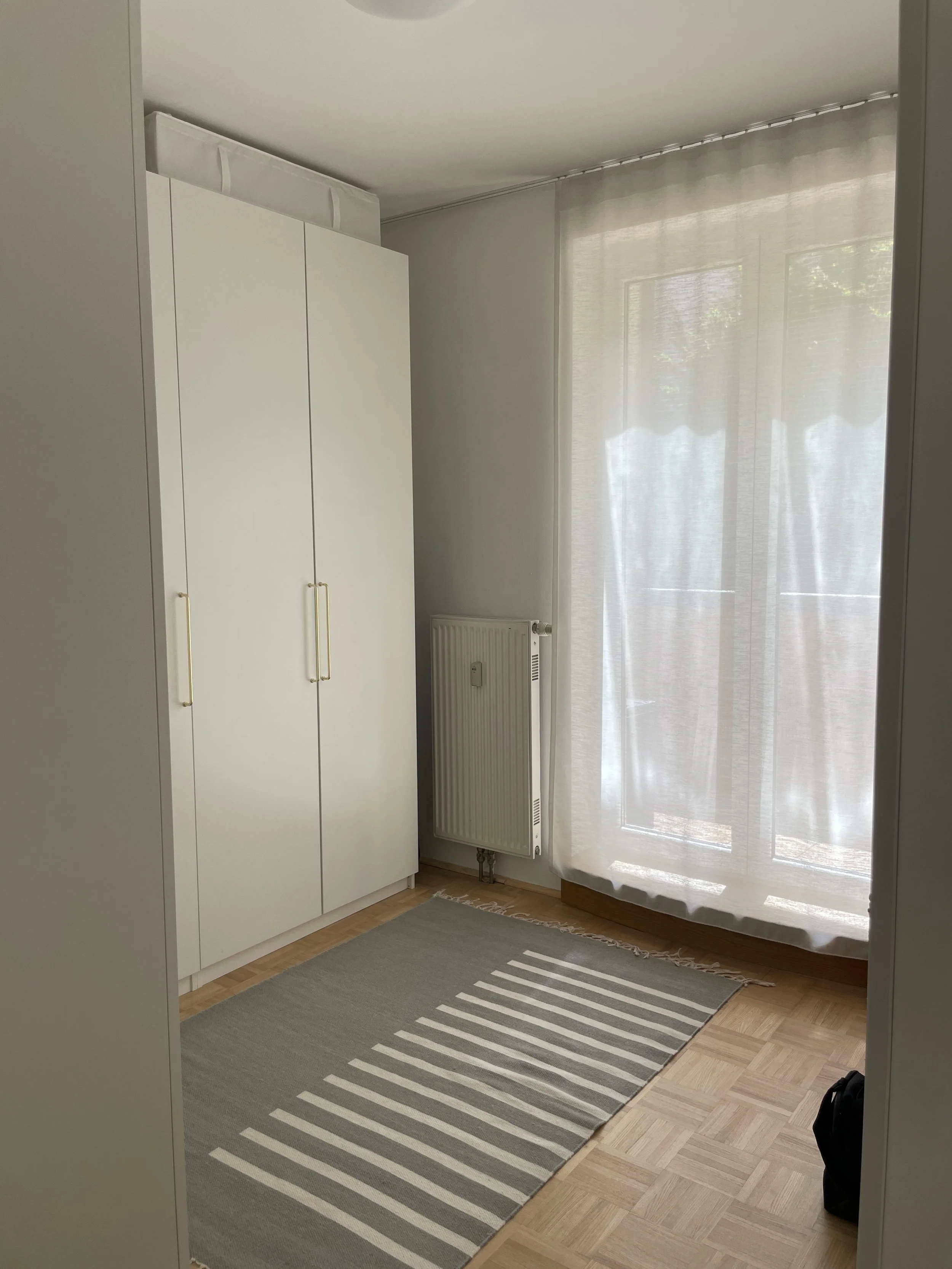 Zimmer mit einem weißen Kleiderschrank, einem grauen gestreiften Teppich und einem Fenster mit lichtdurchlässigem Vorhang.