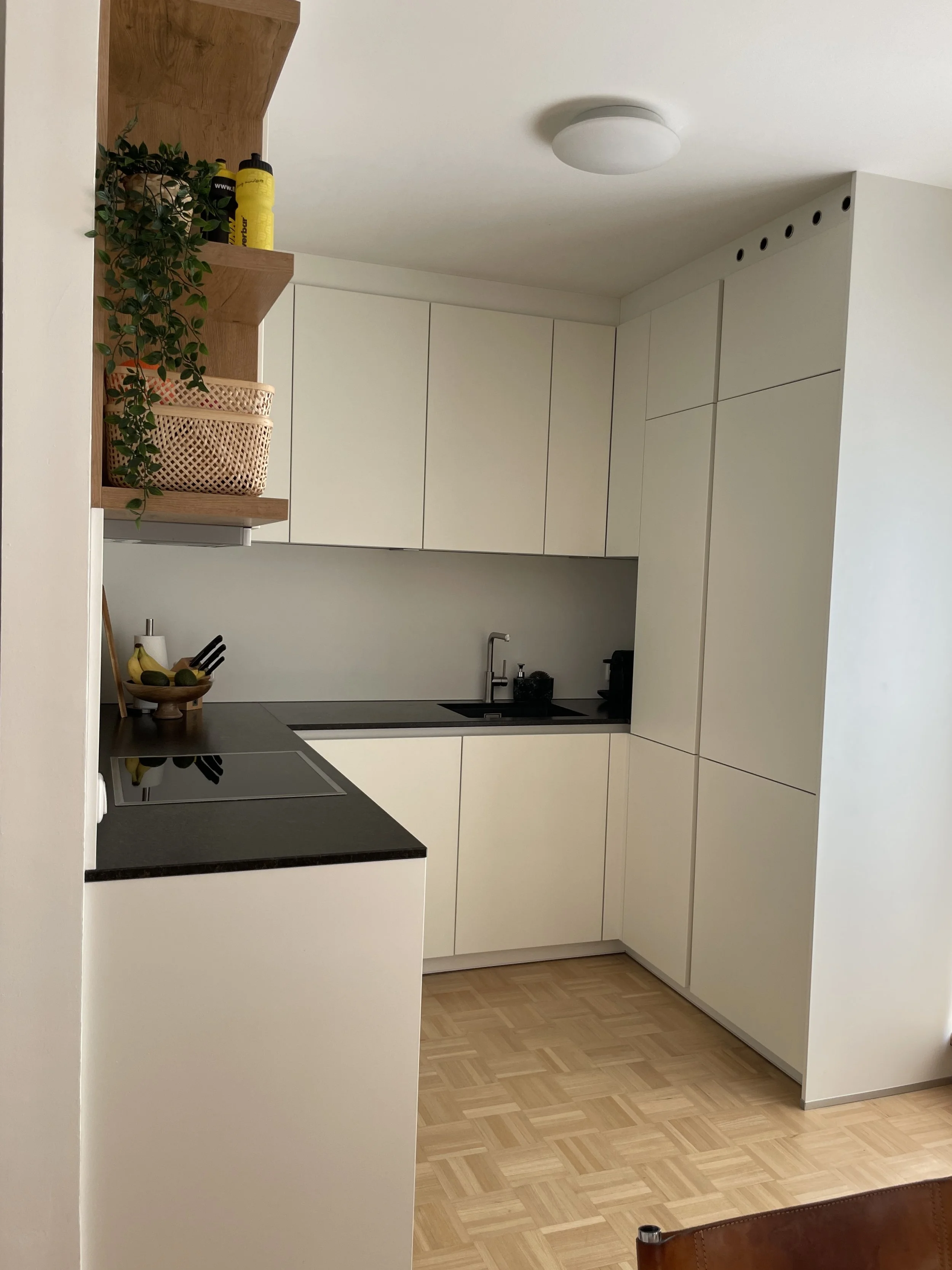 Moderne Küche mit weißen Schränken, schwarzen Arbeitsplatten und einem Elektroherd. Pflanzen und Deko auf Holzregalen, hölzerner Bodenbelag.