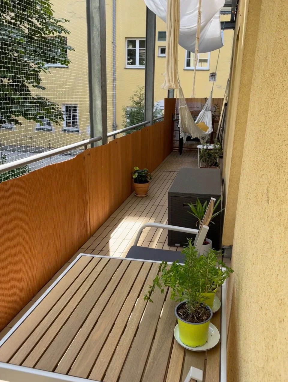 Schmaler Balkon mit Holzverkleidung, Hängematte, Pflanzen in Töpfen und Tisch mit Holzplatte. Gebäude im Hintergrund.