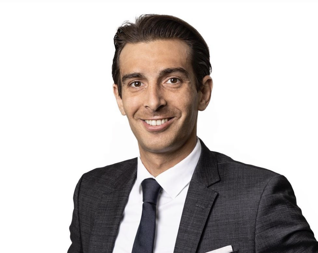 Porträt eines lächelnden Mannes in einem dunkelgrauen Anzug mit weißem Hemd und schwarzer Krawatte vor einem weißen Hintergrund.