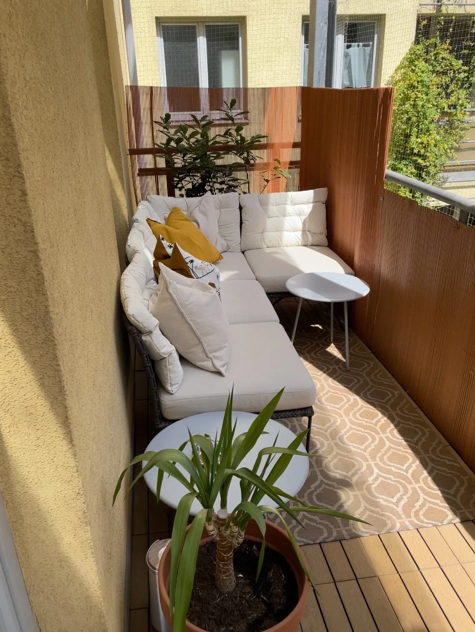 Kleine gepflegte Terrasse mit weißem Ecksofa, gelben und weißen Kissen, kleinem runden Tisch in Weiß, Zimmerpflanze in einem Blumentopf und Holzbodenbelag. Holzzaun zur Privatsphäre.