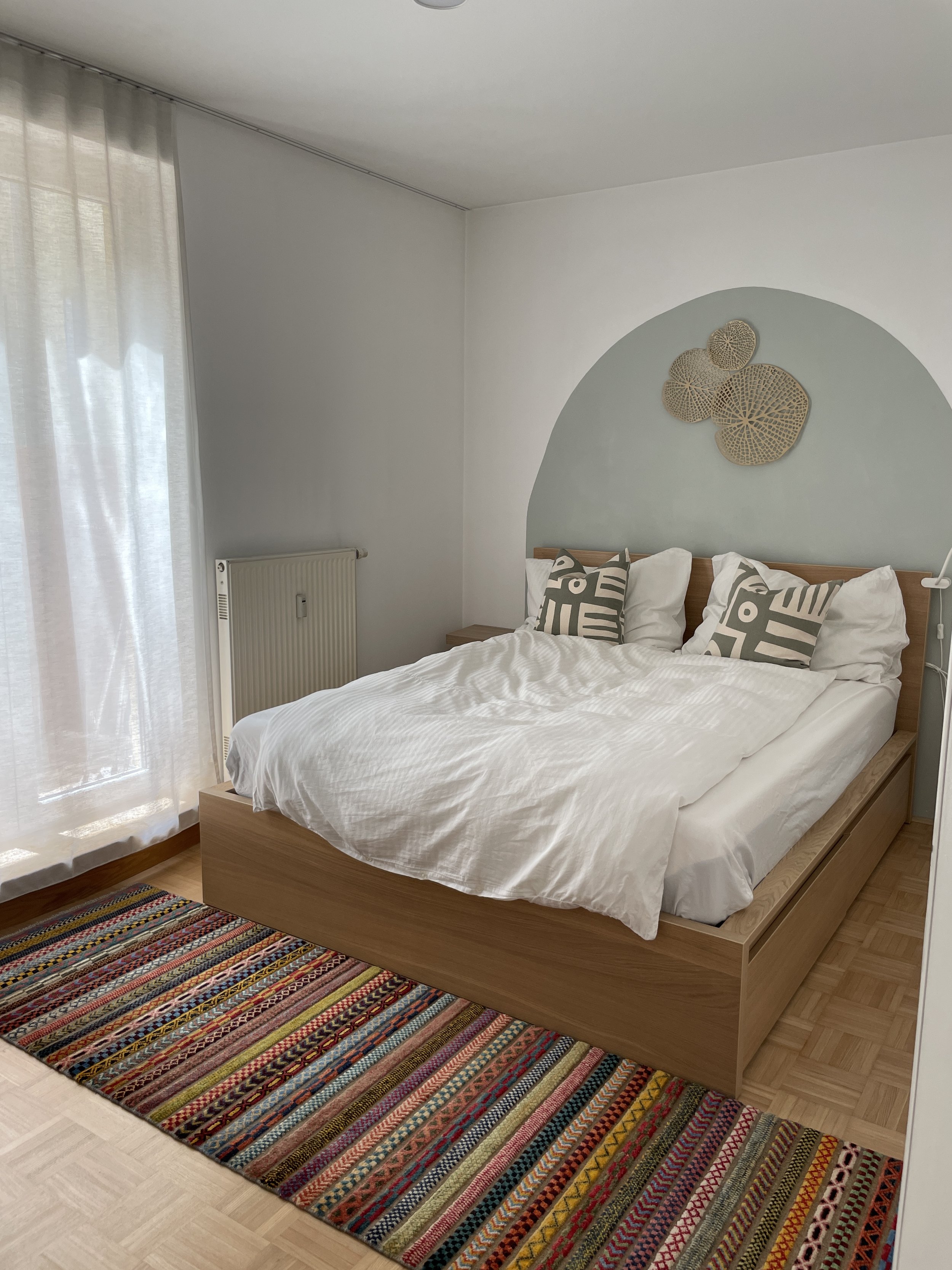 Modernes Schlafzimmer mit einem Holzbett, weißen Bettlaken und geometrischen Kissen. An der Wand hinter dem Bett ist ein dekoratives Wandornament. Ein bunter Teppich liegt auf einem Holzboden. Vorhänge sind an einem Fenster neben dem Bett zu sehen.