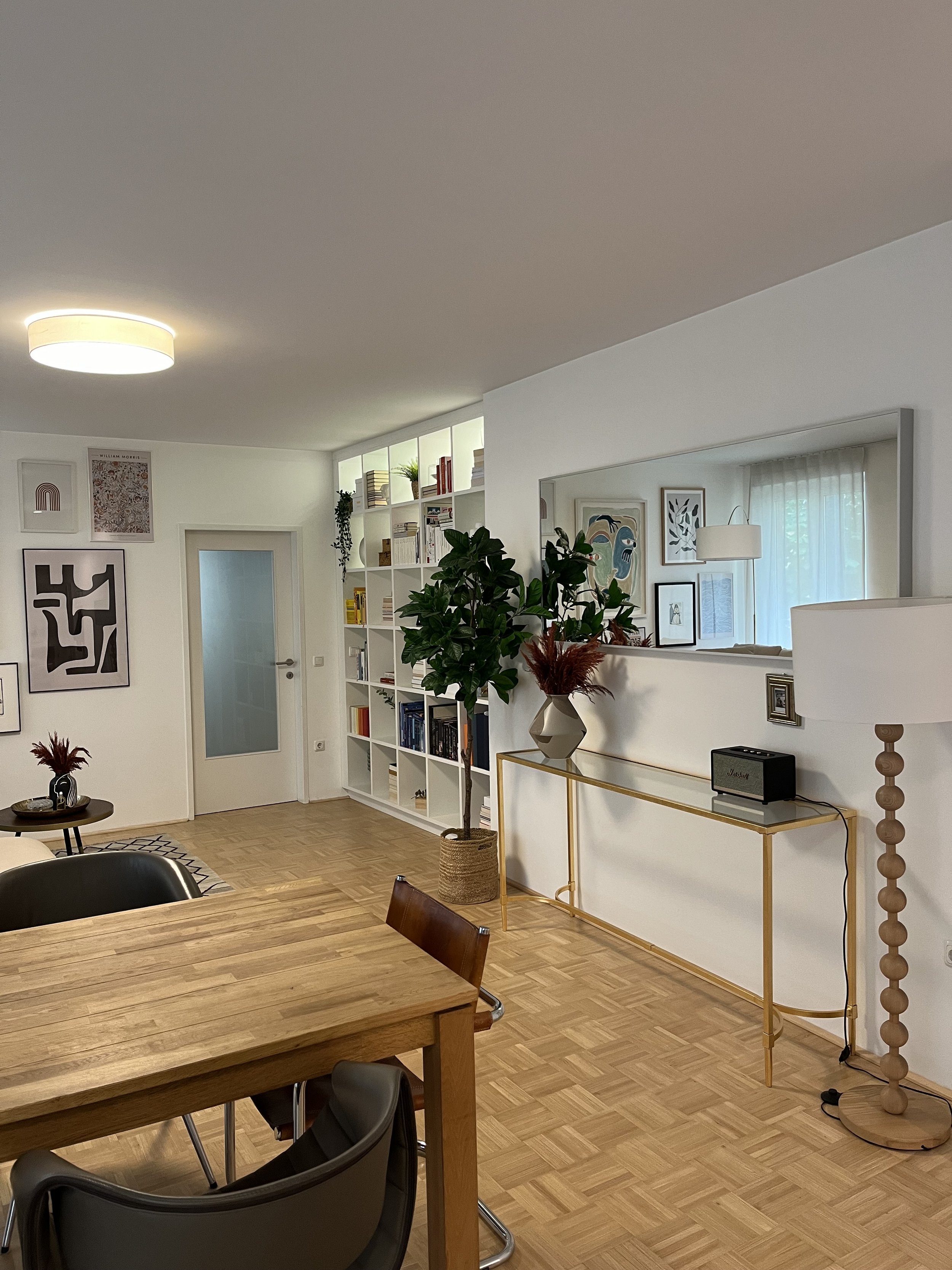 Modernes Wohnzimmer mit Holztisch, Stühlen, Regalen mit Büchern und Pflanzen, dekorativen Bilderrahmen an der Wand, Stehlampe, Spiegel und Lautsprecher.