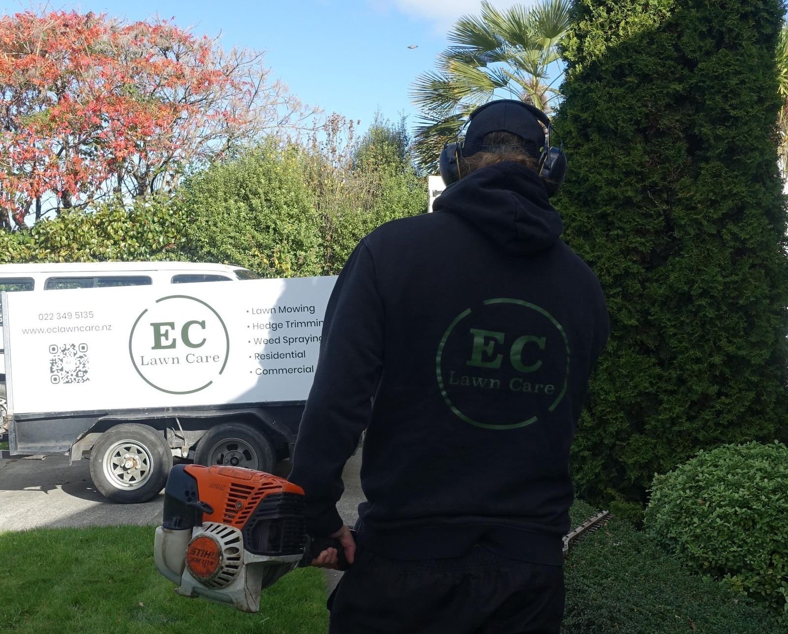 EC Lawn Care operator wearing branded workwear and carrying a STIHL hedge trimmer beside the company trailer at a residential property in Hawke's Bay.