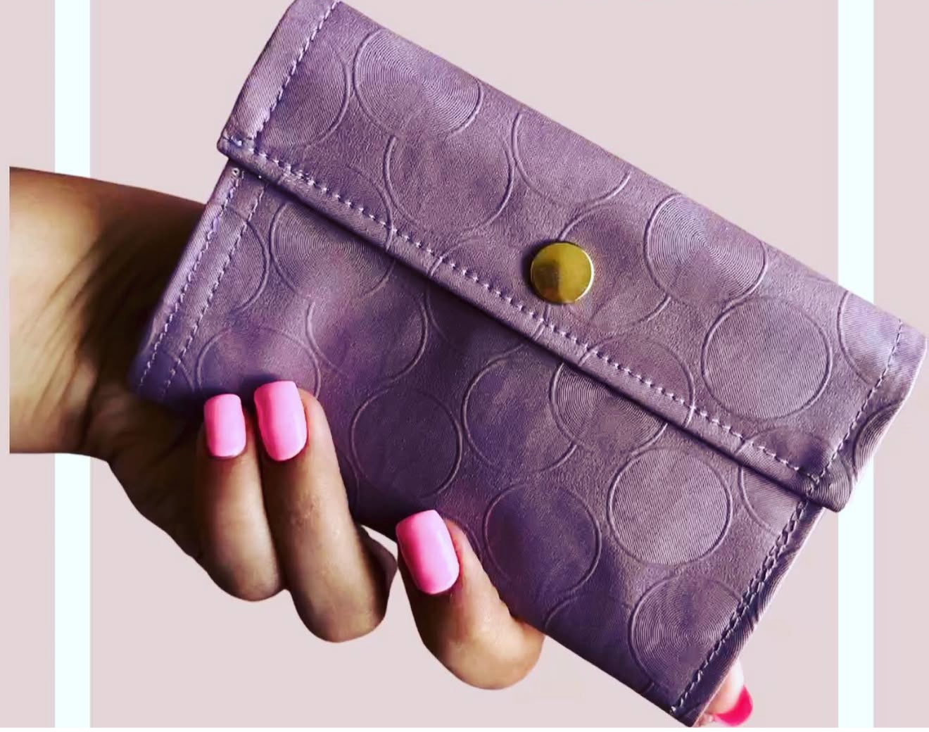 A purple clutch purse with a circular pattern, held by a hand with pink nail polish, against a light pink background.