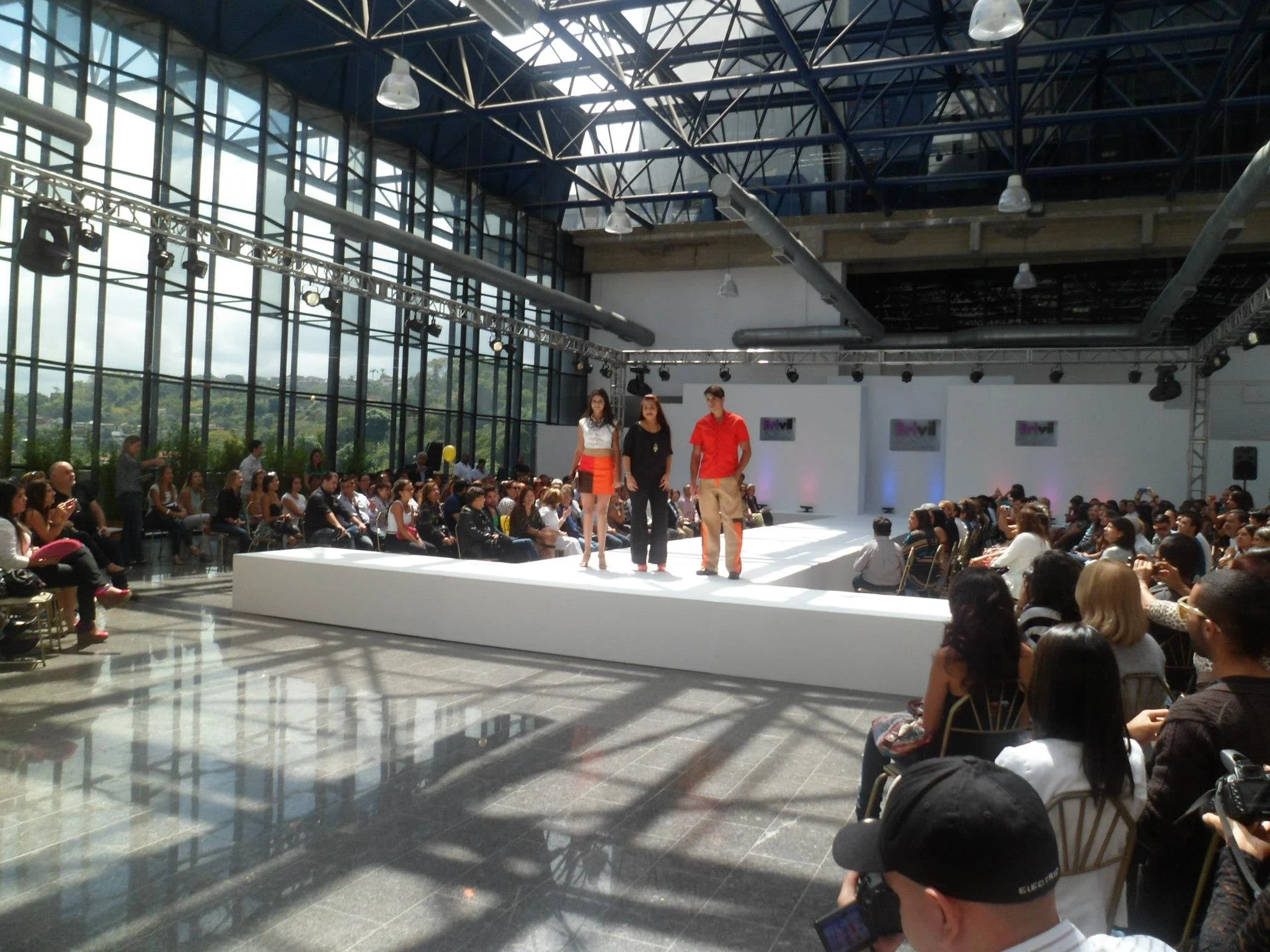 Fashion models walk on a runway stage during a fashion show inside a large, glass-walled venue with an audience seated on either side.