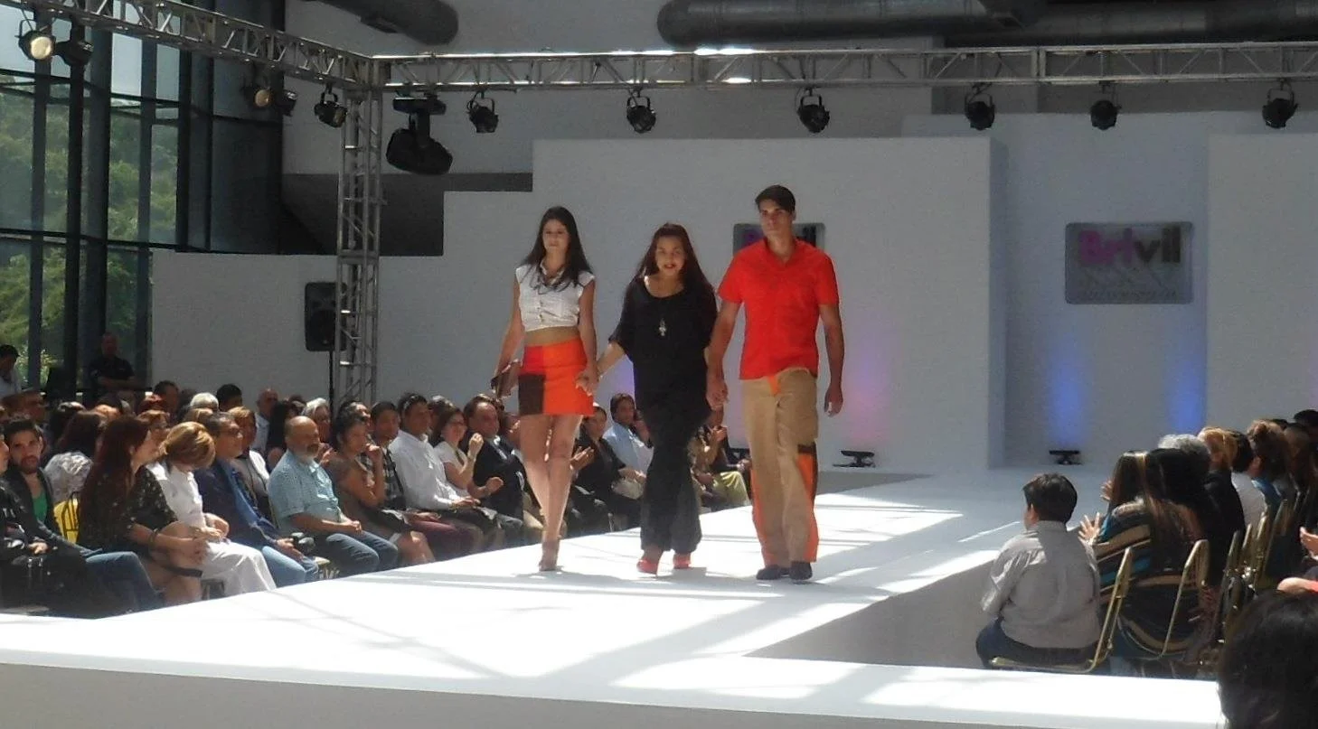 Three young models walking on a stage runway during a fashion show, with audience seated on both sides and large windows in the background.