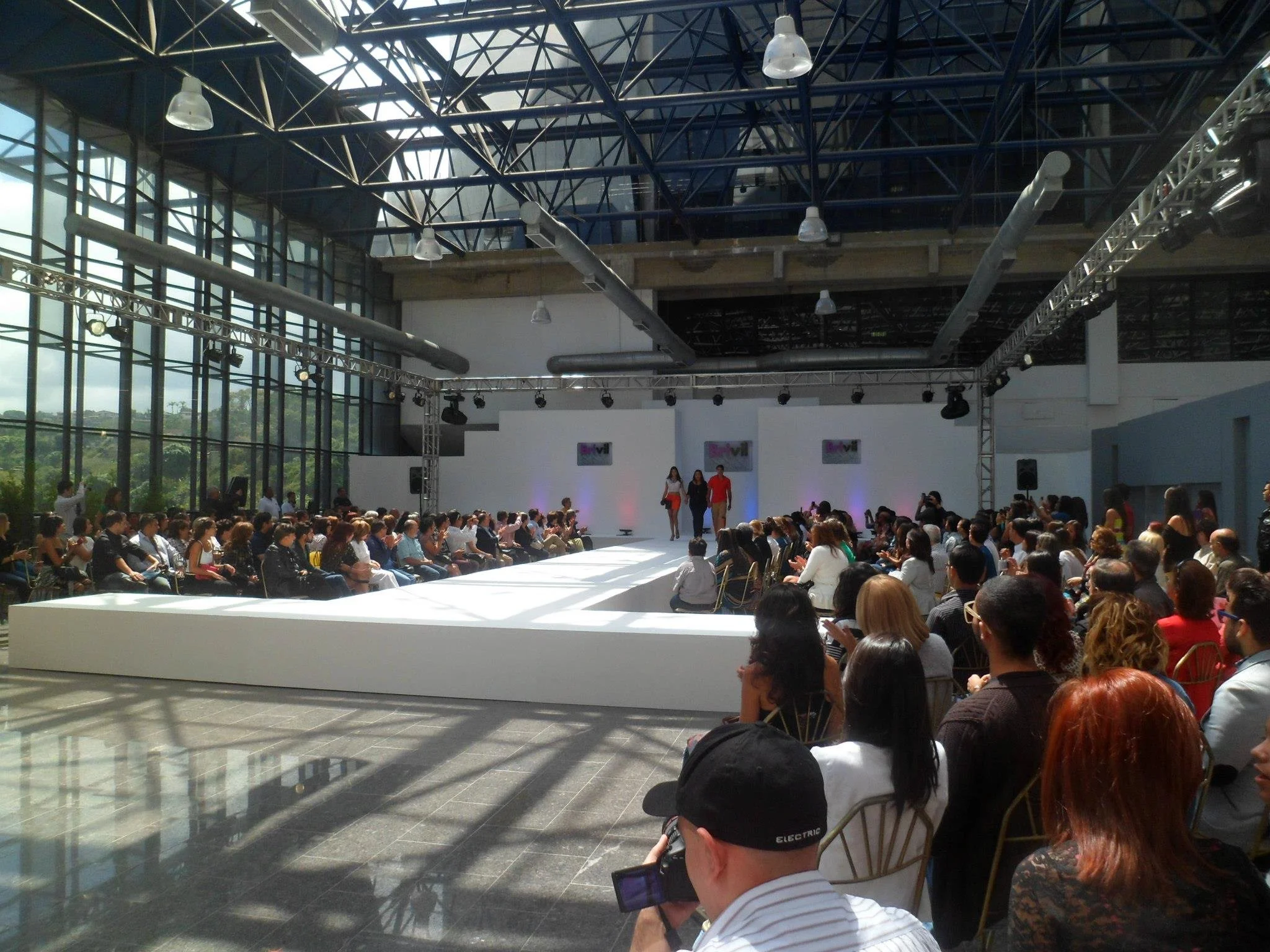 Fashion show runway inside a large glass-walled venue with an audience seated on both sides, and models walking on the runway.