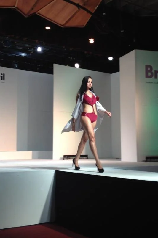 A woman walking on a runway during a fashion show, wearing red lingerie with a partly open silver robe, black high heels, and long dark hair.
