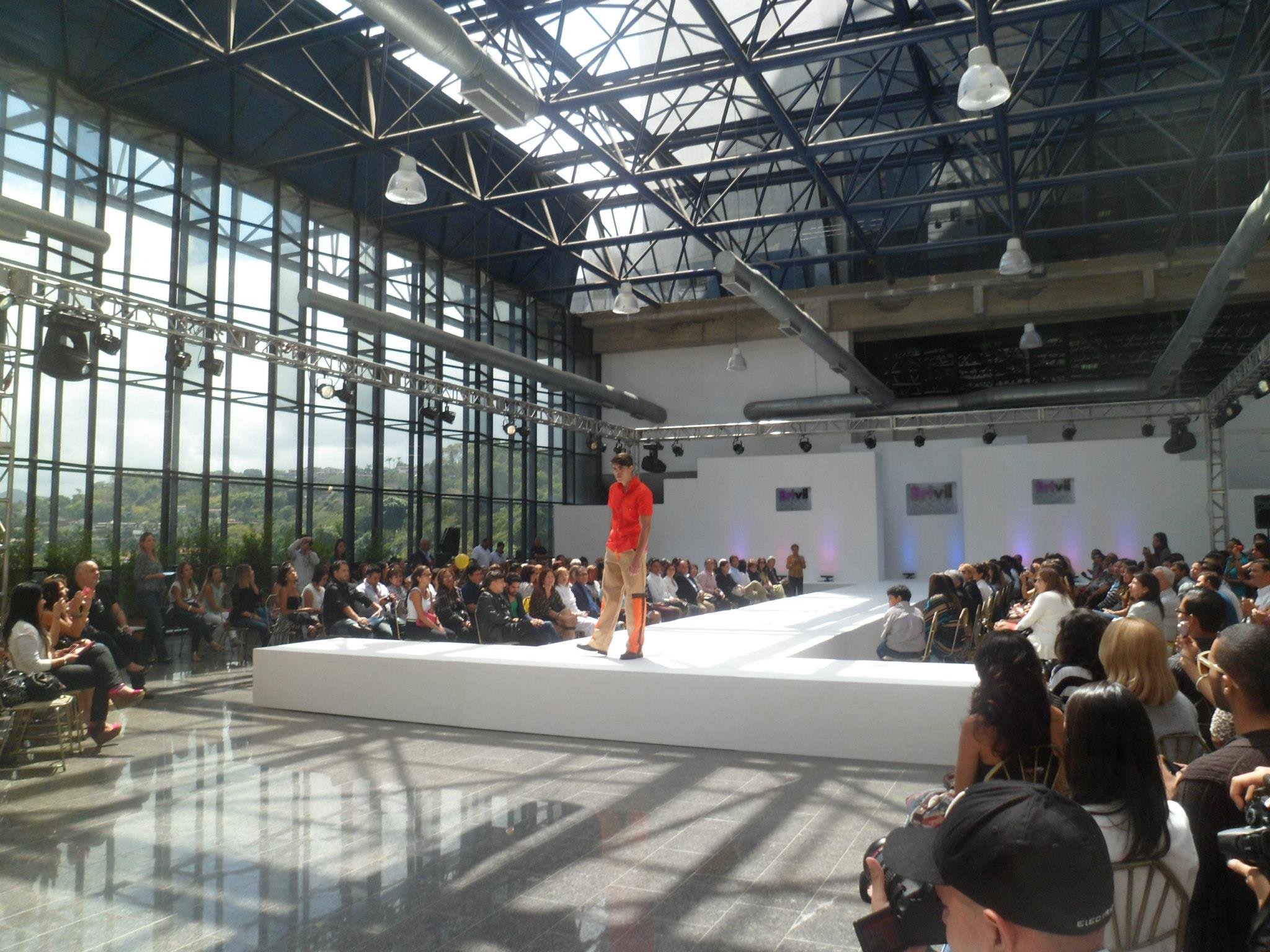 Fashion show runway in a large glass-walled venue with a male model walking on the runway, surrounded by seated audience members during daytime.