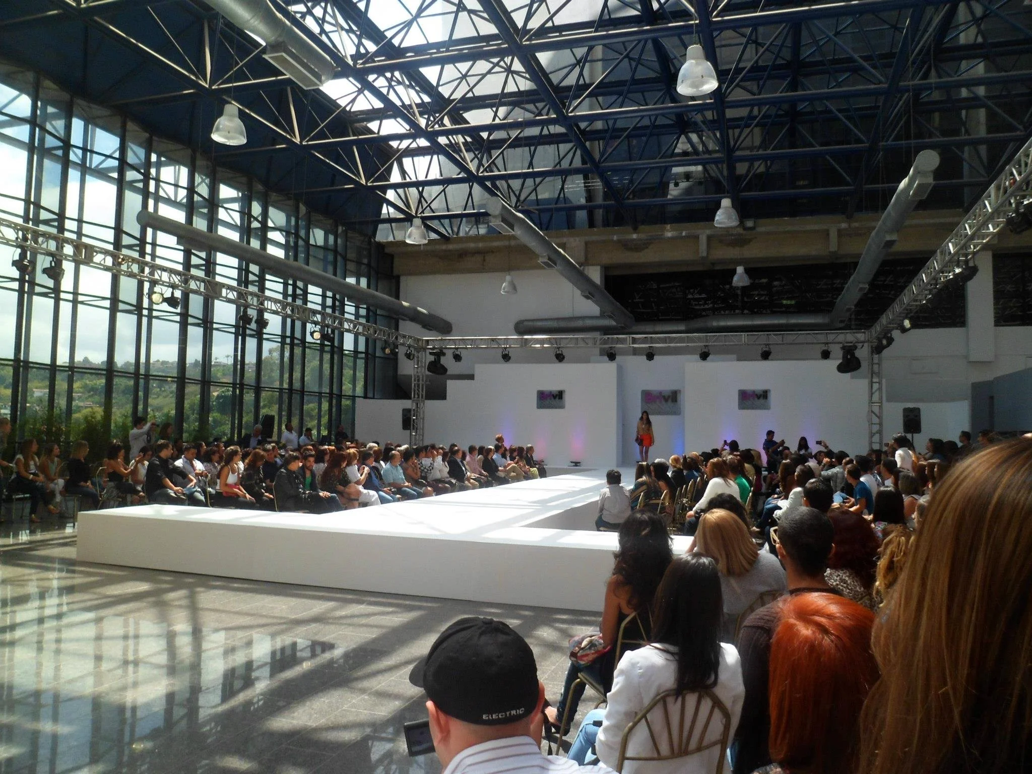 Large indoor event with a runway in the center and many audience members seated on both sides. There are large windows on the left letting in natural light, and a stage at the back with two screens displaying the word 'Revil' and a woman standing on 