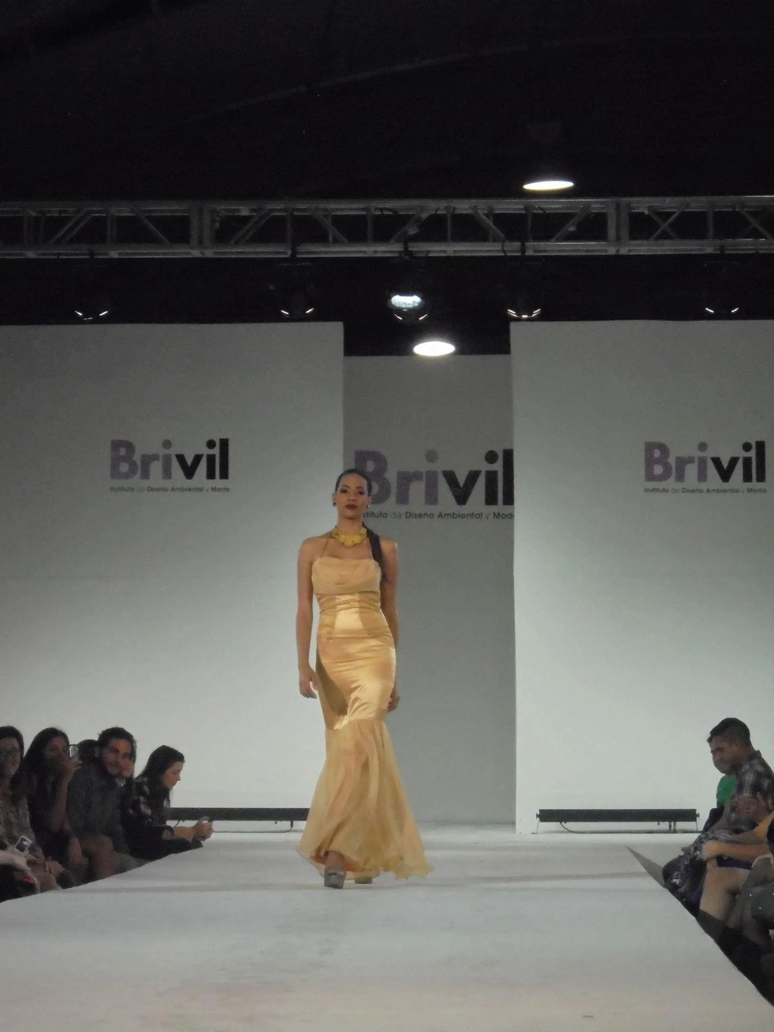 Model walking down runway at fashion show with audience seated on both sides, backdrop displaying 'Brivil' branding, bright lighting overhead.