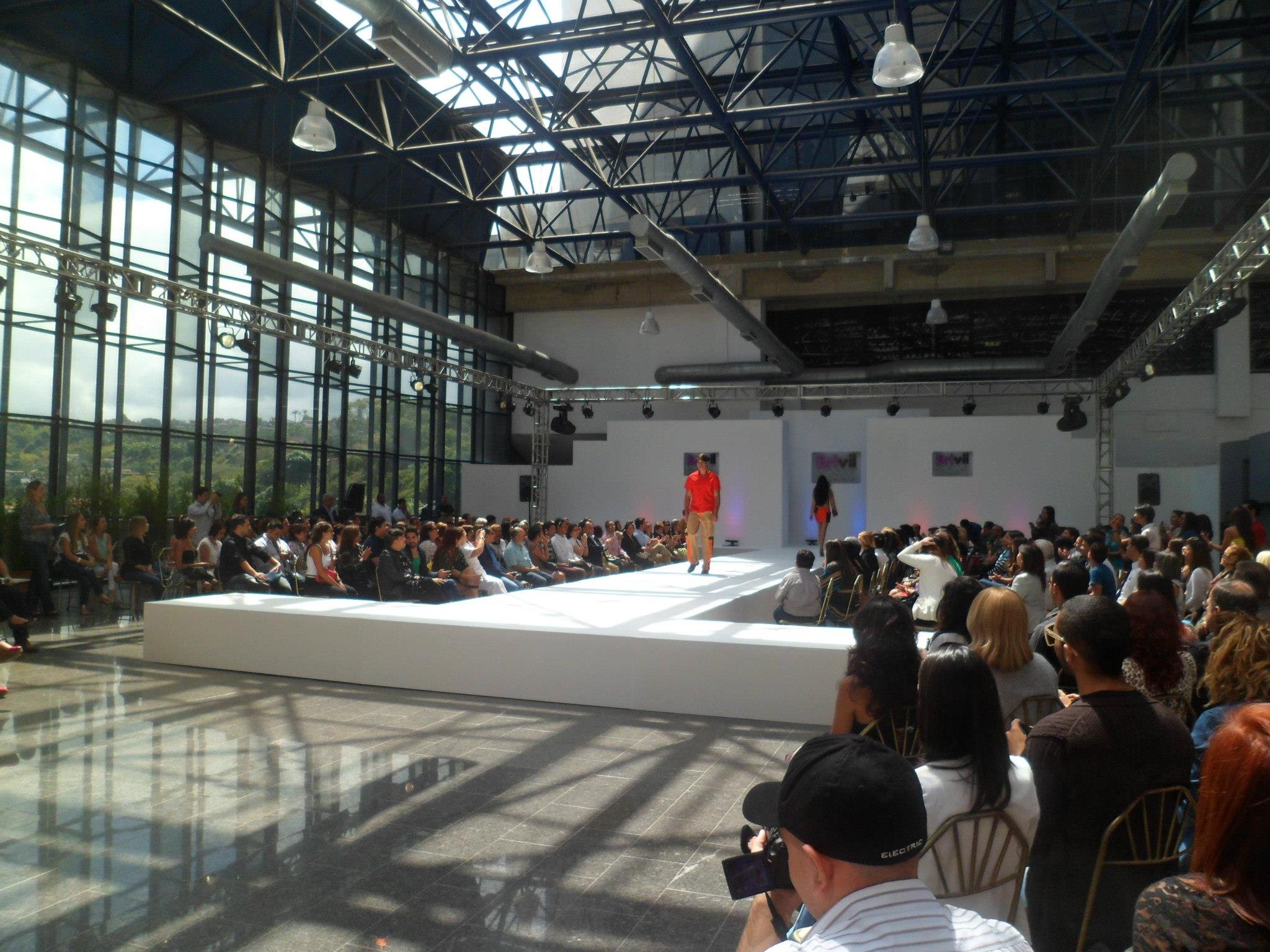Model walking on a runway during a fashion show in a spacious, modern venue with large glass windows and high ceiling.