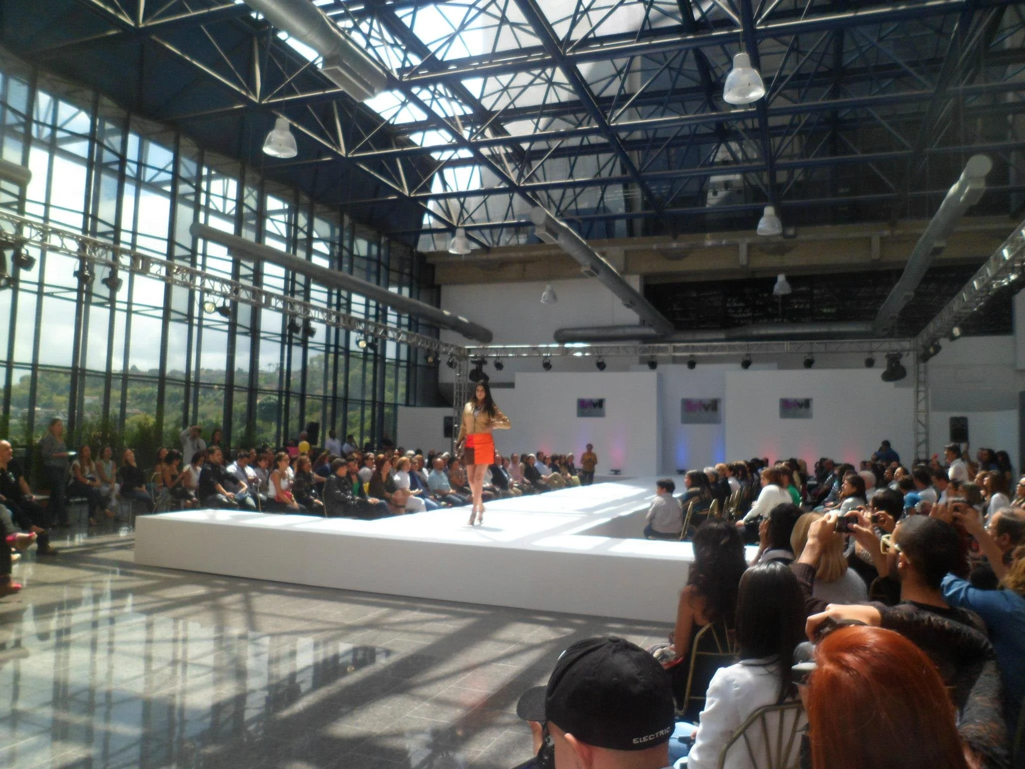 Fashion runway show in a large, glass-walled industrial-style venue with audience seated on both sides, model walking on the runway.