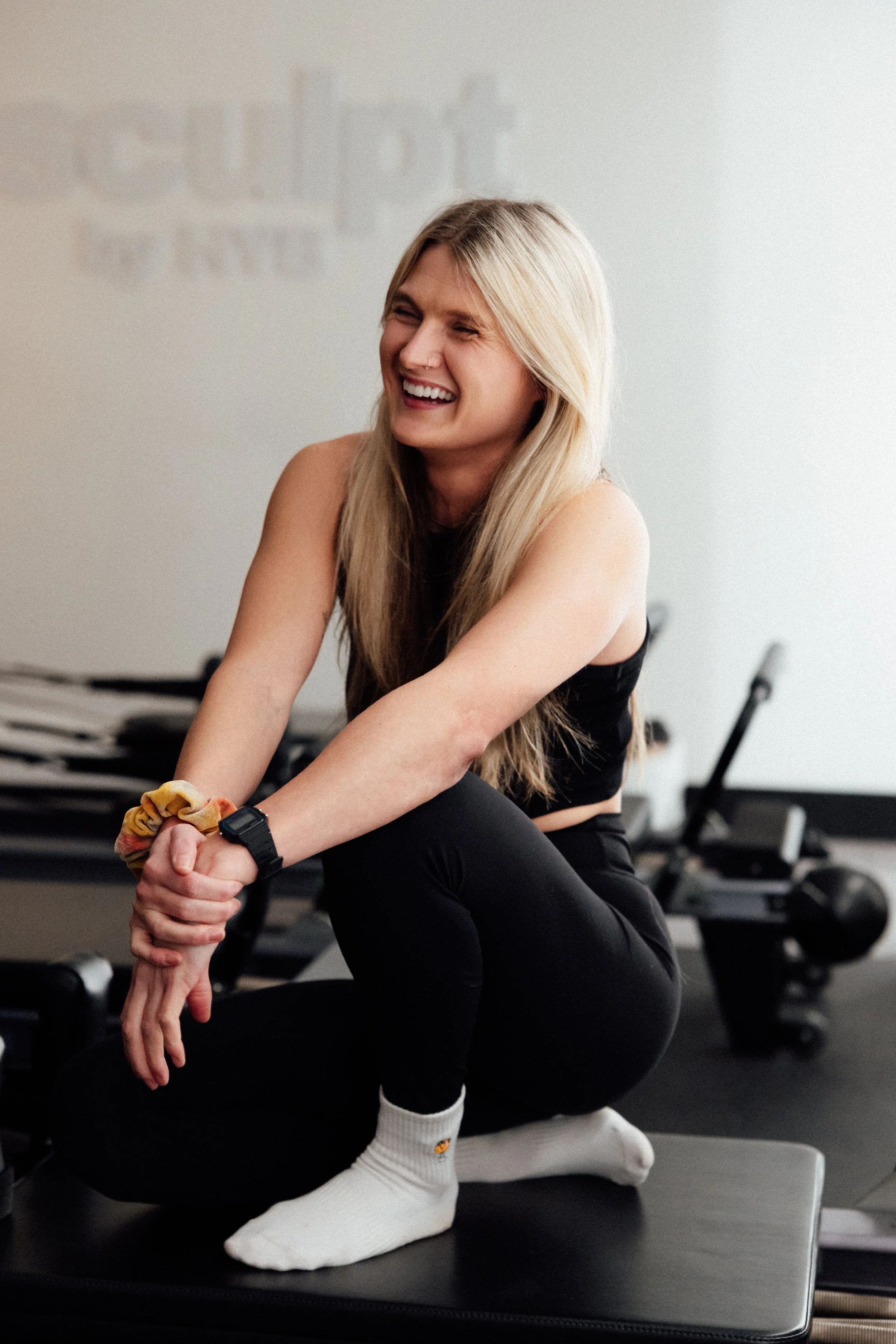 A young woman with blonde hair smiling and sitting on a black exercise mat in a gym, dressed in black workout clothes and white socks.