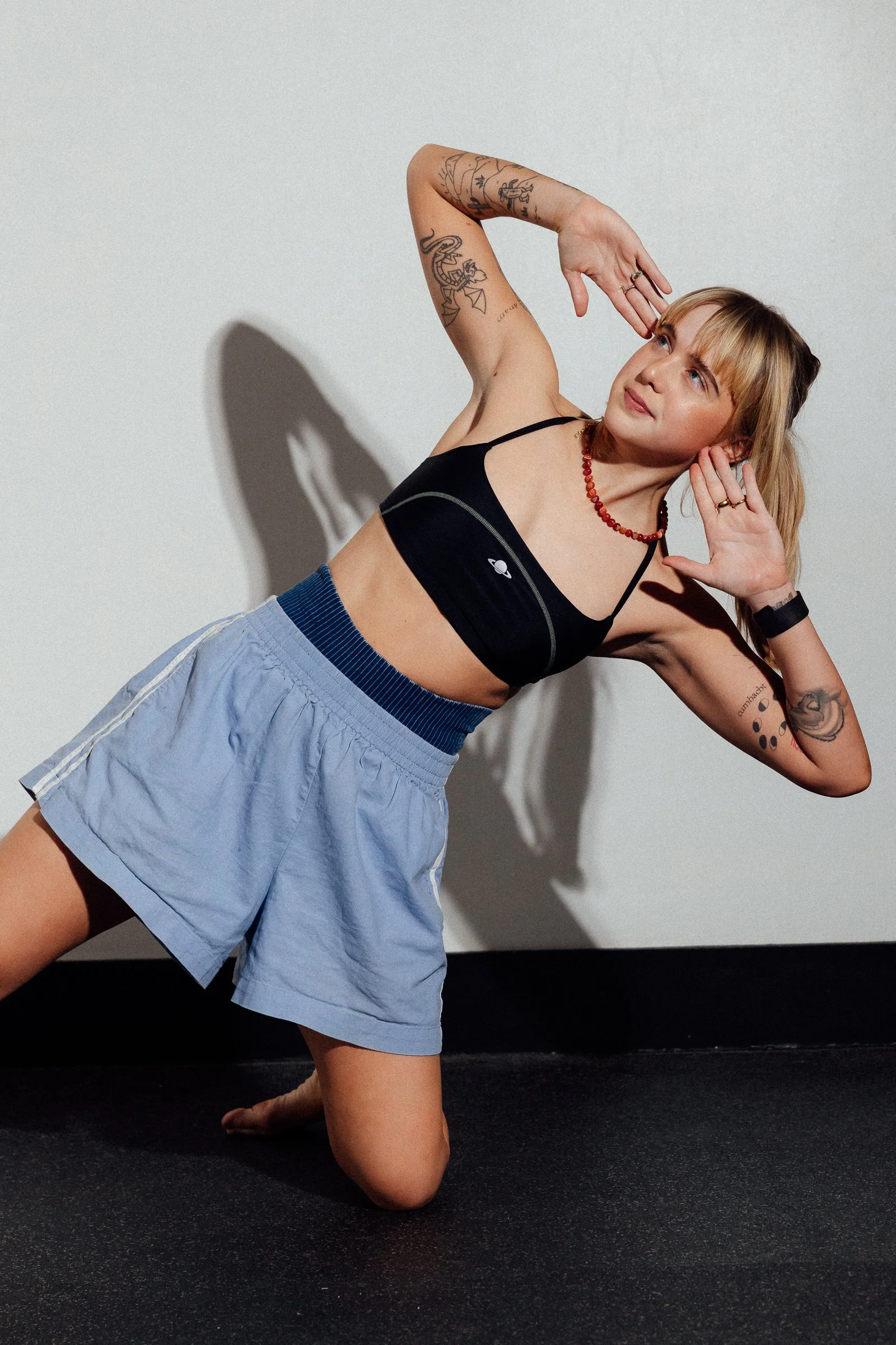 A young woman with tattoos, wearing a black sports bra, light blue shorts, and a red necklace, poses in a yoga stretch on a black floor against a white wall.