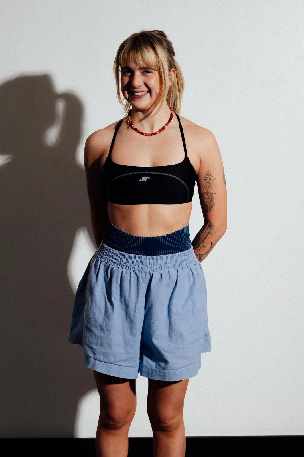 A young woman with blonde hair, tattoos on her arms, and a red beaded necklace, wearing a black crop top with a planet logo and light blue shorts, smiling and standing against a plain white wall.