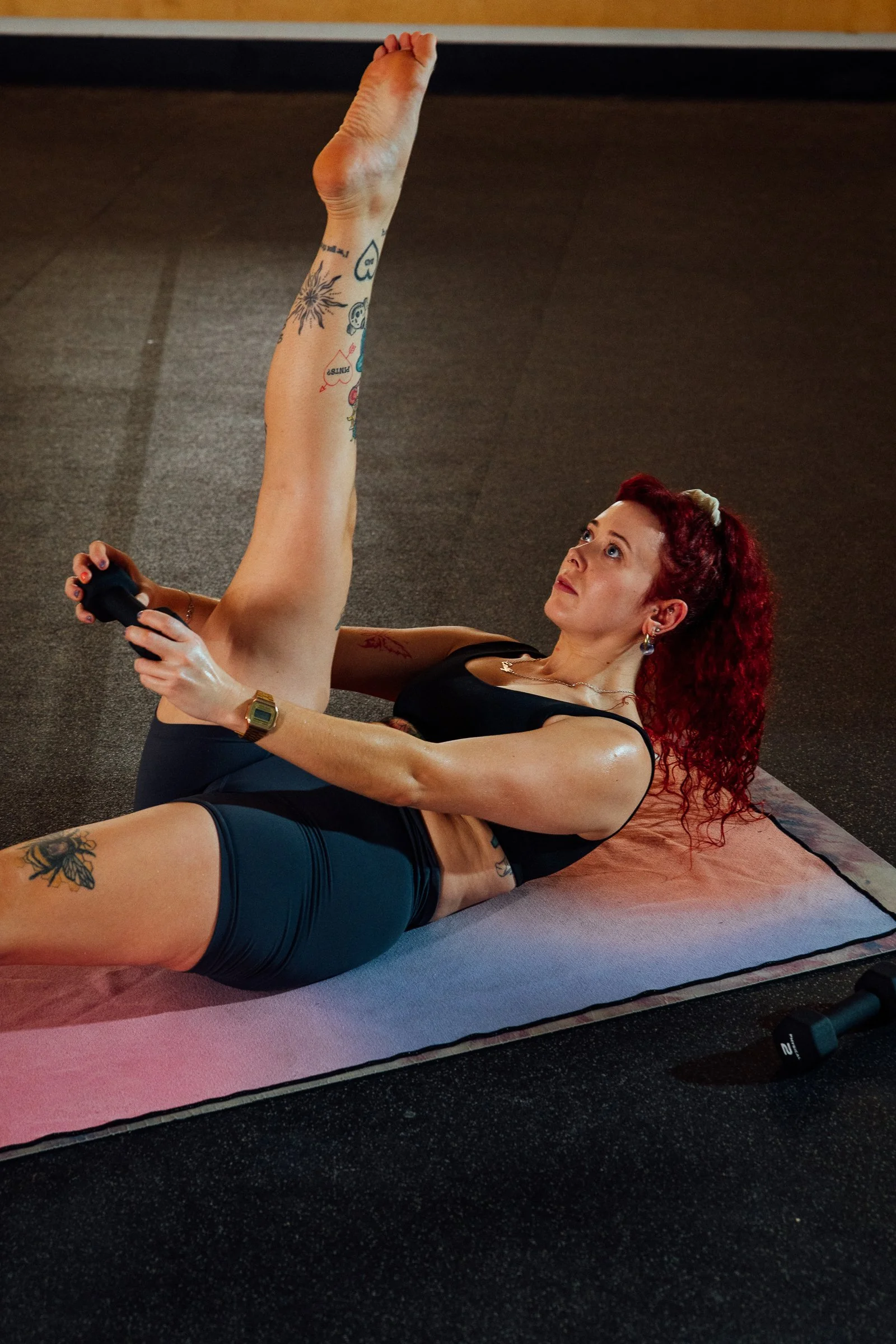 A woman with curly red hair performs a fitness exercise on a yoga mat inside a gym, holding a dumbbell and extending one leg upward.