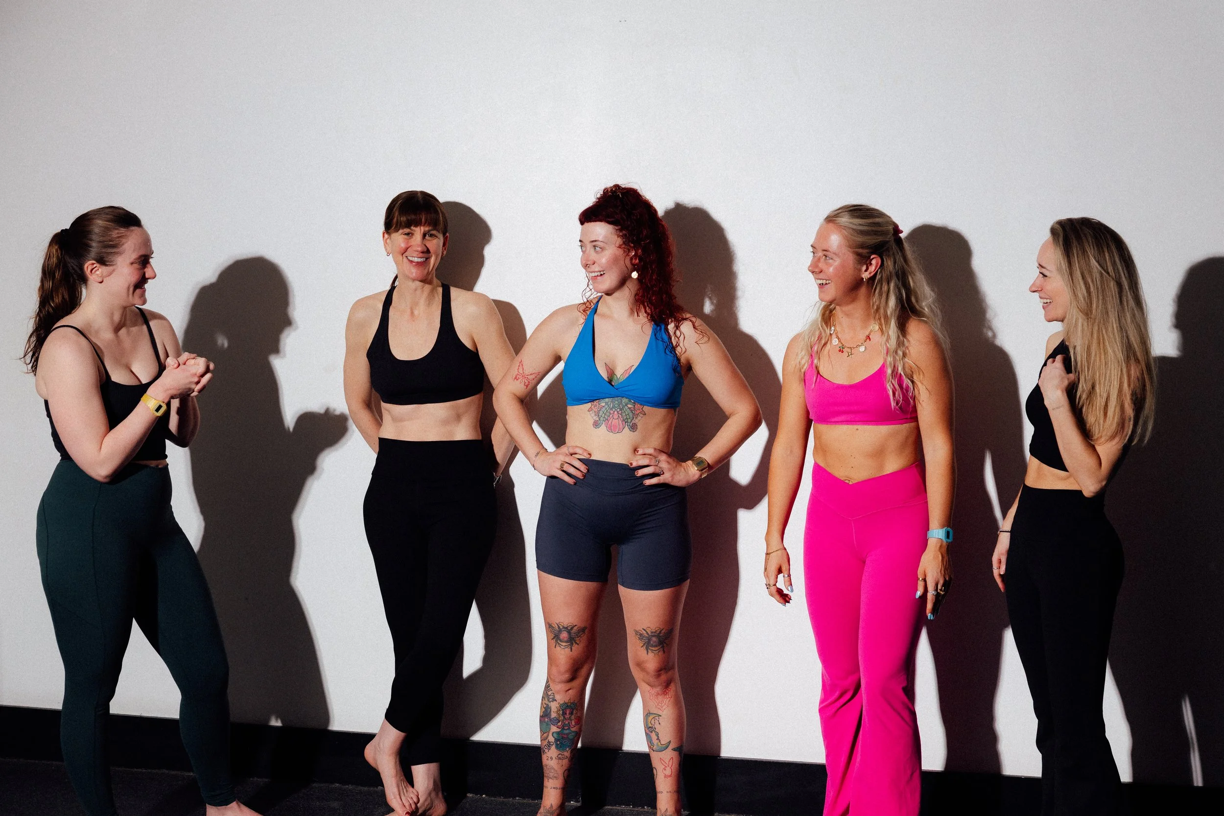 Five women standing against a plain white wall, smiling and engaging in conversation, with shadows cast behind them.