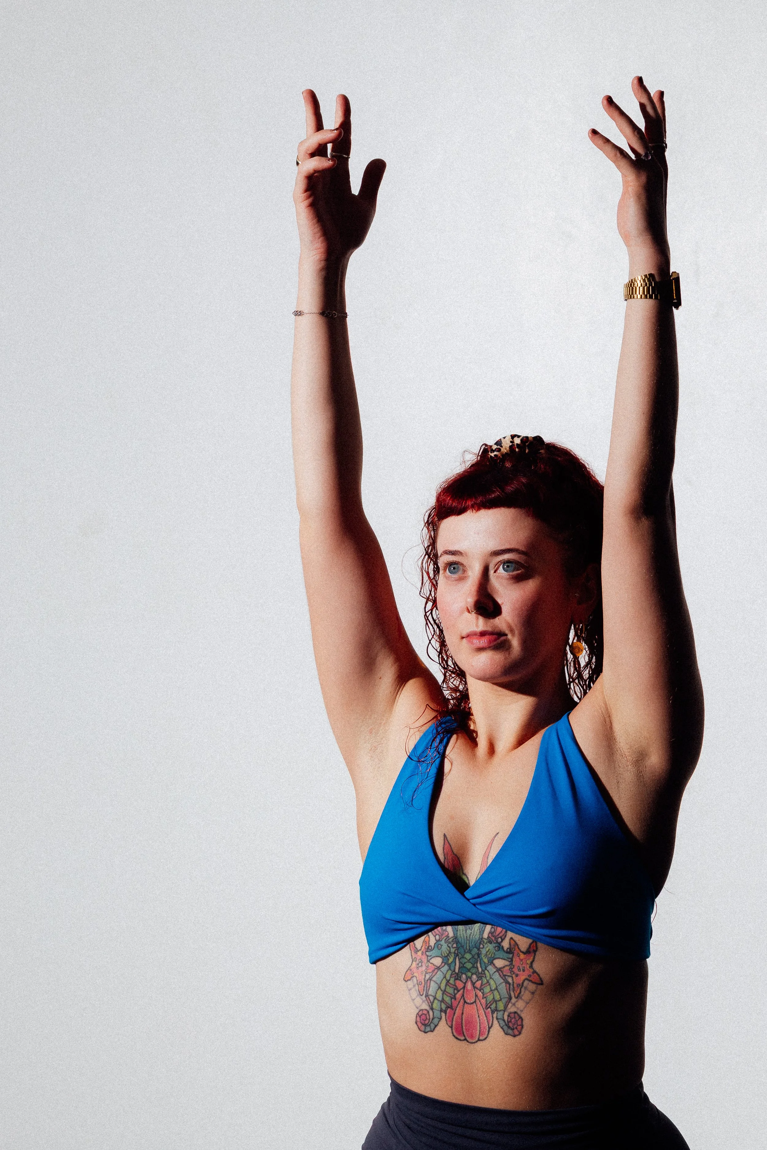 A woman with red hair and tattoos wears a blue crop top and has her arms raised.