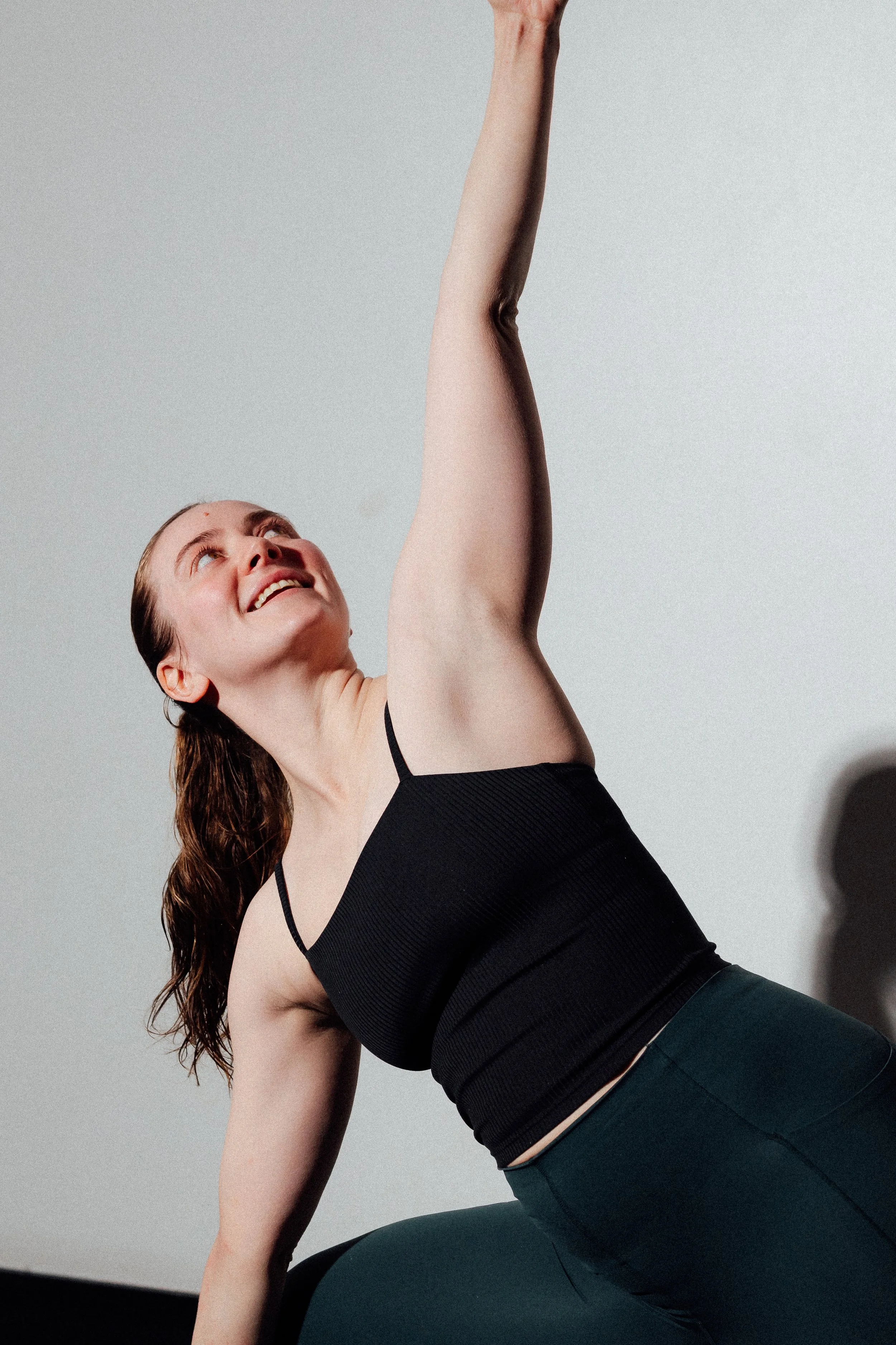 A woman with brown hair in a black sports top and dark leggings performing a yoga pose on a black mat in a bright room.