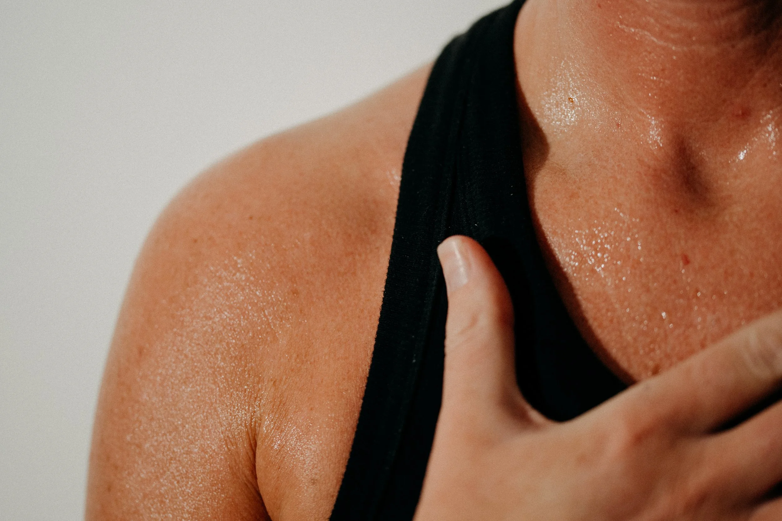 Close-up of a woman with sweat on her shoulder and neck, wearing a black tank top, touching her shoulder with her right hand.