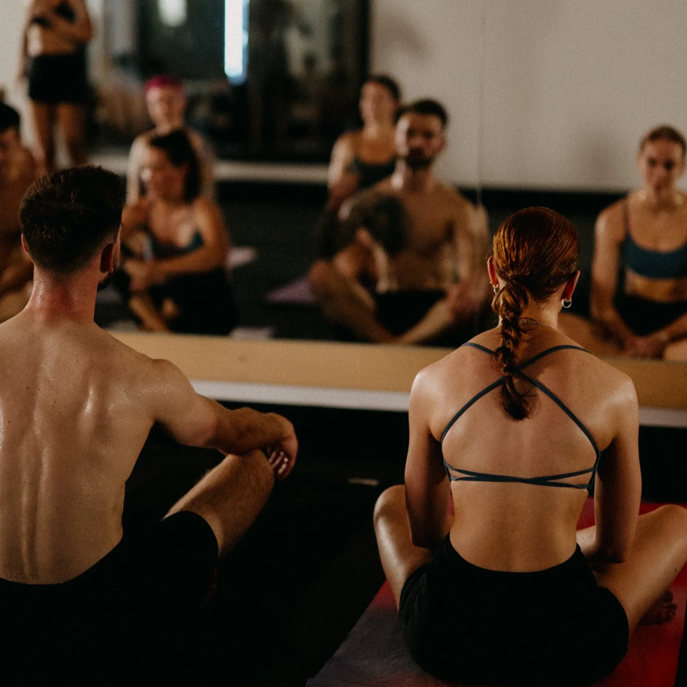 Group of people participating in a yoga class, sitting on mats and facing a mirror, with one person leading the session.