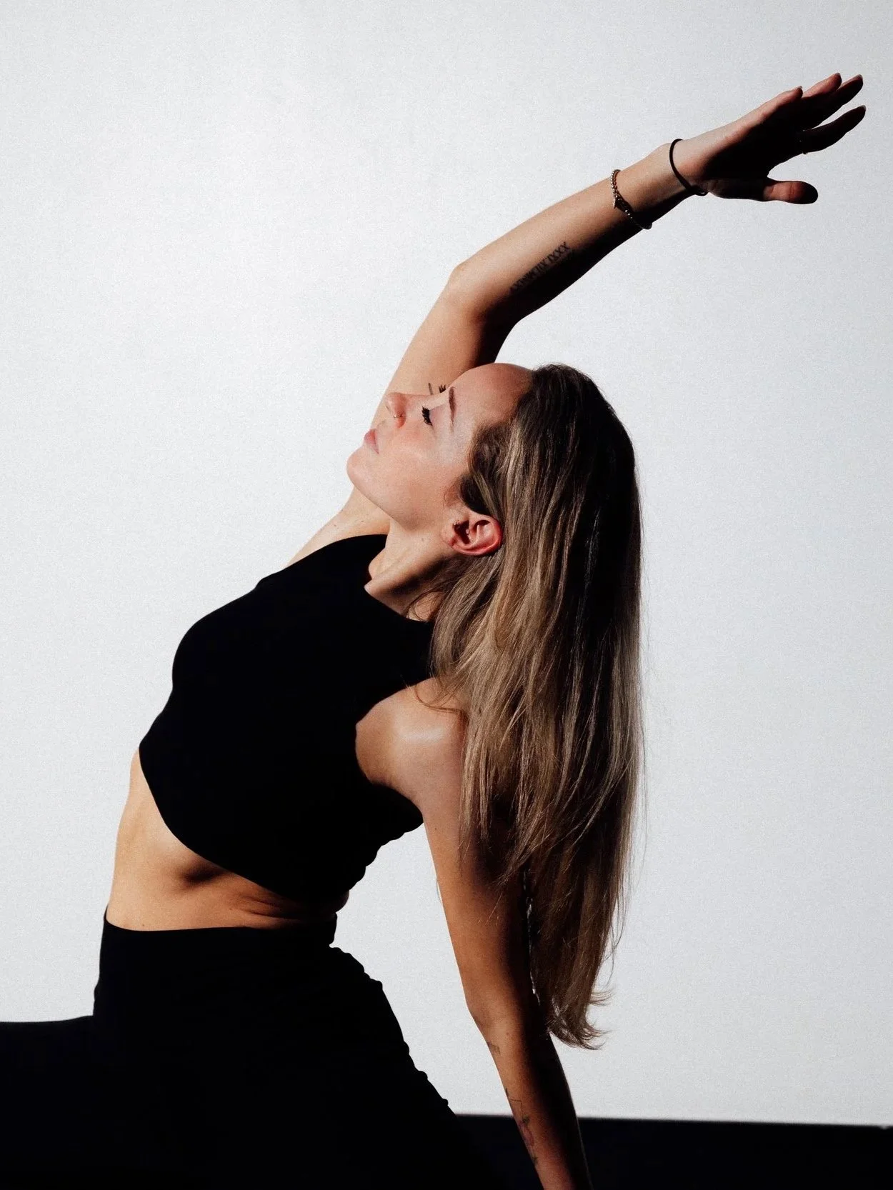 A woman with long brown hair in a black outfit is performing a yoga pose against a plain white background. She is sitting with one leg bent and her upper body leaning back, with her arm extended upward and her face turned slightly to the side.