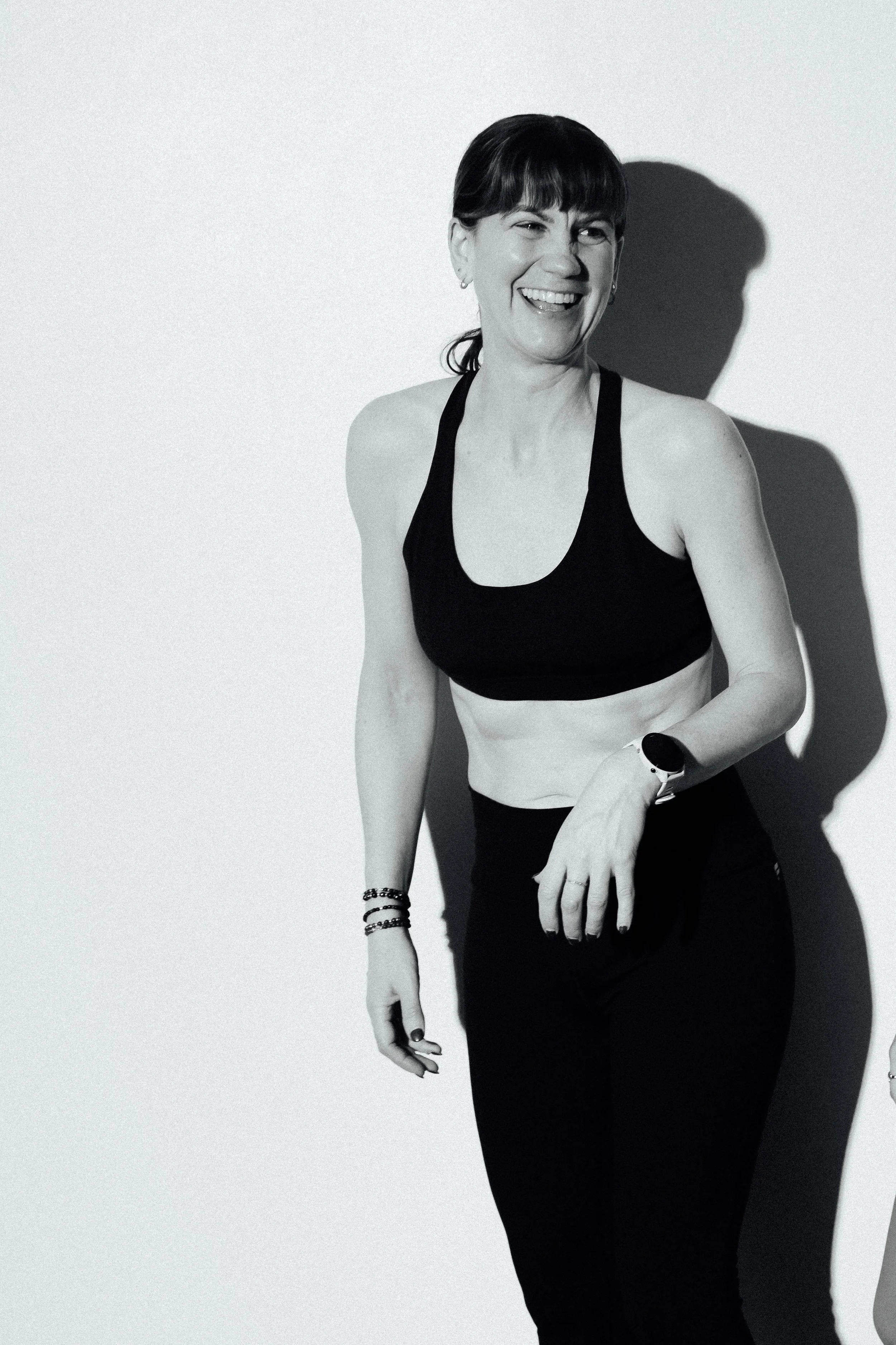 A woman smiling and laughing, wearing workout clothes and a smartwatch, standing against a plain wall with a shadow behind her.