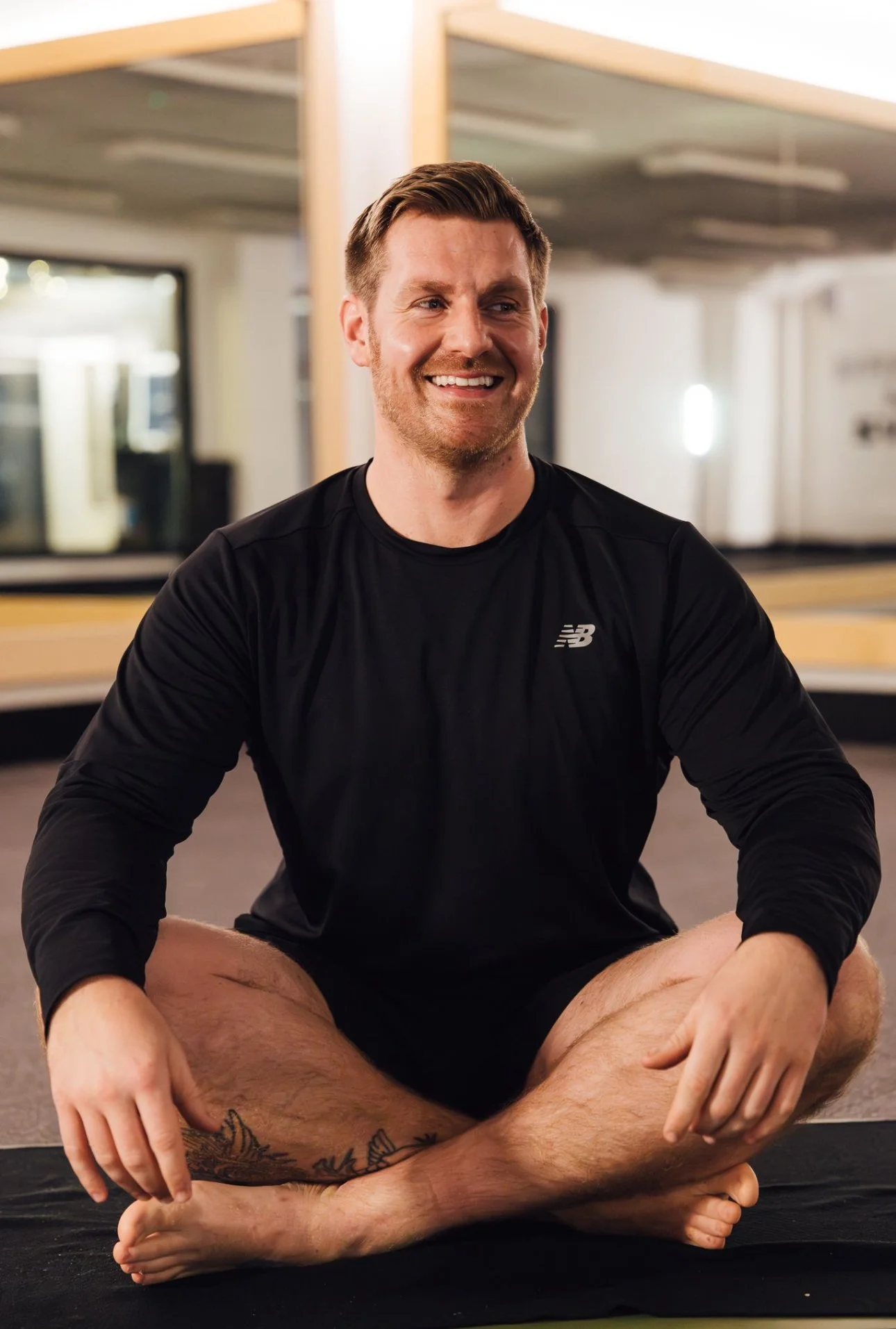 A man sitting cross-legged on a yoga mat in a gym, smiling, wearing a black athletic shirt, with mirrors and gym equipment in the background.