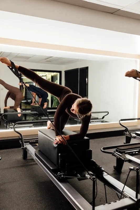 A person in a black workout outfit performing a Pilates exercise on a reformer machine in a Pilates studio, with mirrors and other exercise equipment visible.