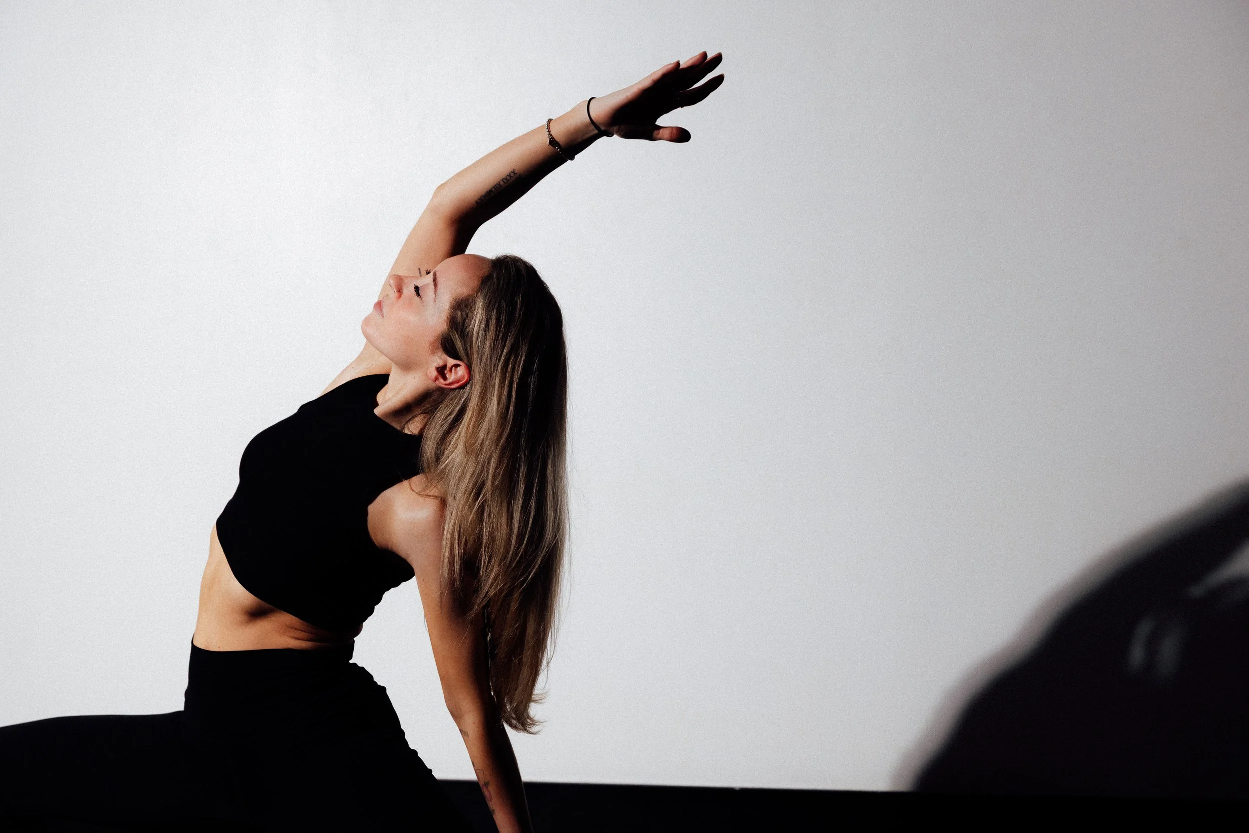 A woman with long hair in a black crop top and black pants performs a yoga pose against a plain gray background, with her left arm extended overhead and her head tilted back.