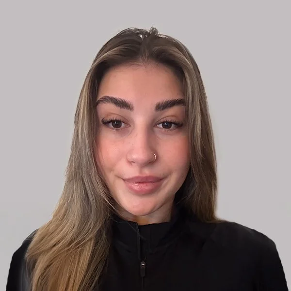 Young woman with long blonde hair, a nose piercing, and a black zip-up jacket, standing against a plain gray background.