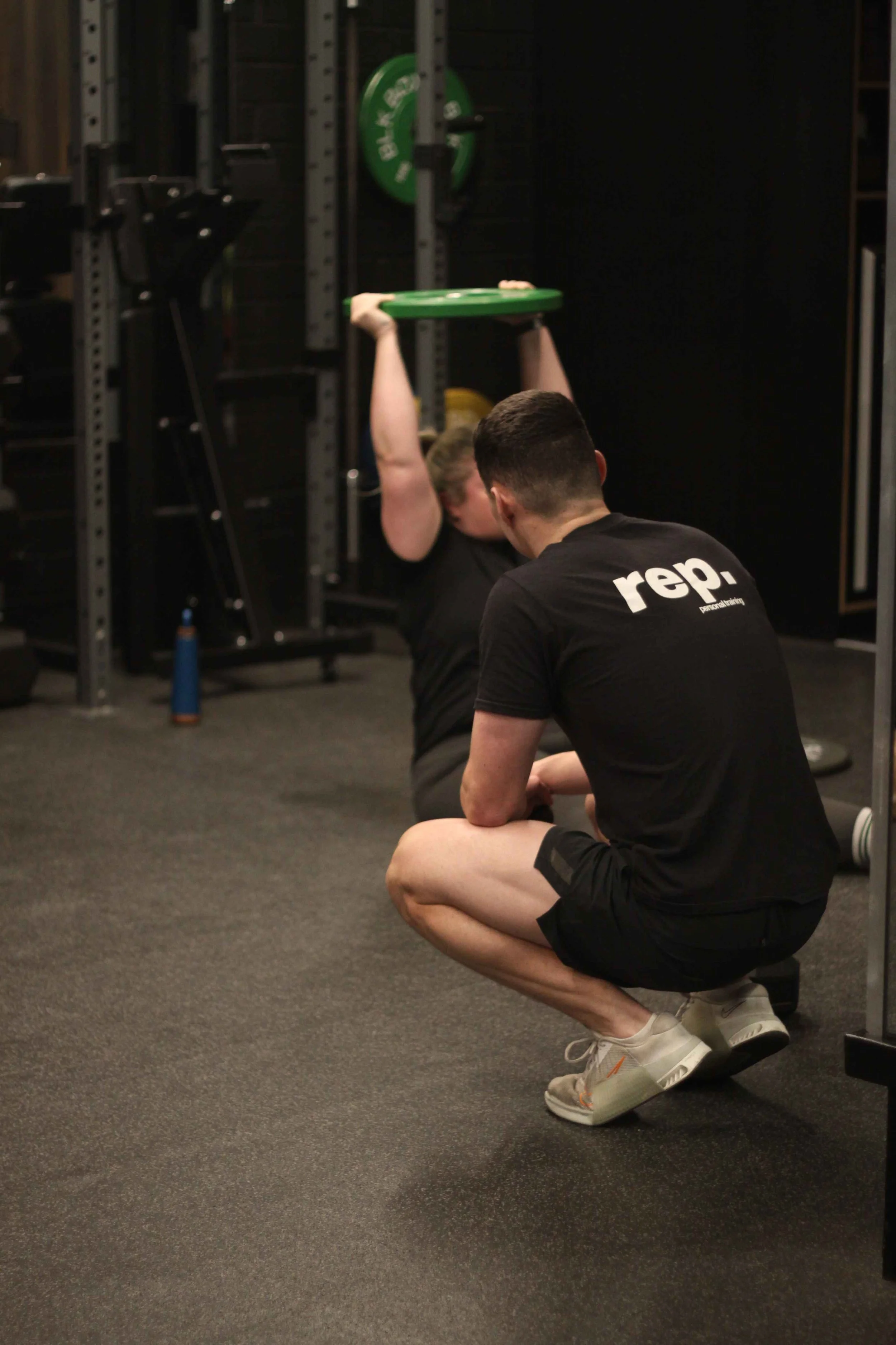 A person in black workout clothes doing an overhead press with a green weight plate, while another person, crouching in front and wearing a black shirt with white print, observes in a gym.