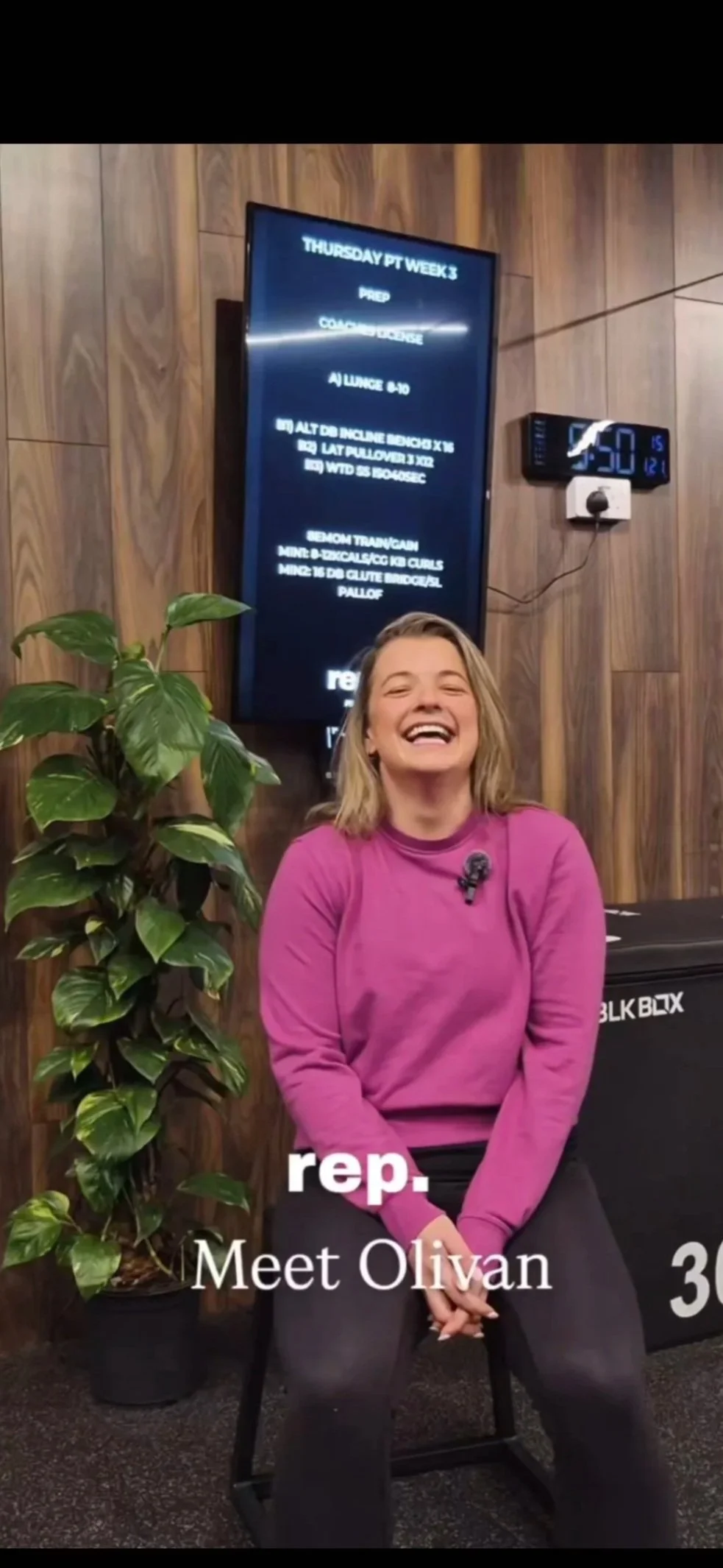 A woman sitting and smiling, wearing a pink sweatshirt and black pants, next to a large leafy plant, with a wooden-paneled wall and a digital schedule display in the background.