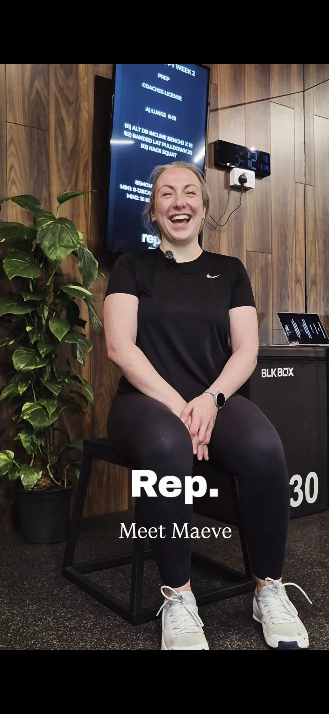 A woman with blonde hair smiling and sitting on a black chair in a gym or fitness studio, wearing a black Nike t-shirt, black leggings, white sneakers, and a smartwatch. Behind her is a wooden wall, a plant to her left, and a digital clock and screen displaying workout details above her. Text on the image reads: "Rep. 30 Meet Maeve."