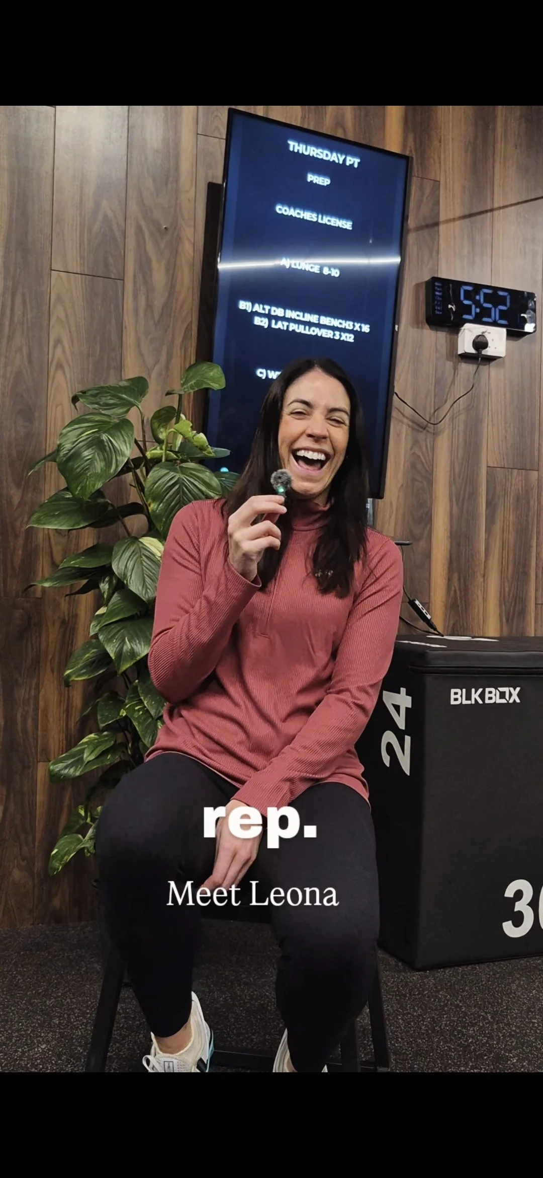 A woman sitting on a chair, smiling and holding a small object, next to a green plant and a black storage box. Behind her is a large digital screen displaying a workout schedule, and a black clock showing the time 5:52.