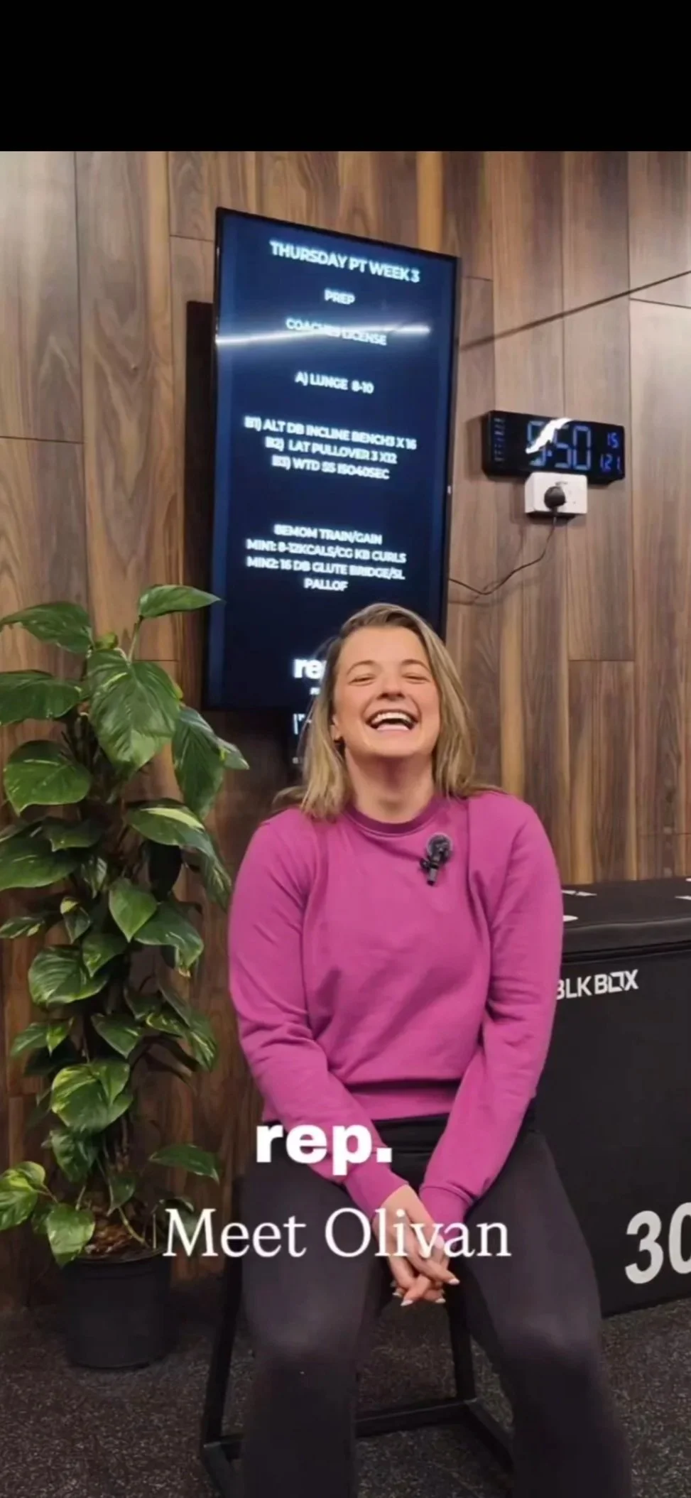A woman in a pink sweatshirt and black pants sitting on a chair indoors, smiling. Behind her, there is a large leaf plant on the left and a digital clock on the wall showing 4:50. A digital display board is mounted on the wall with fitness class details.