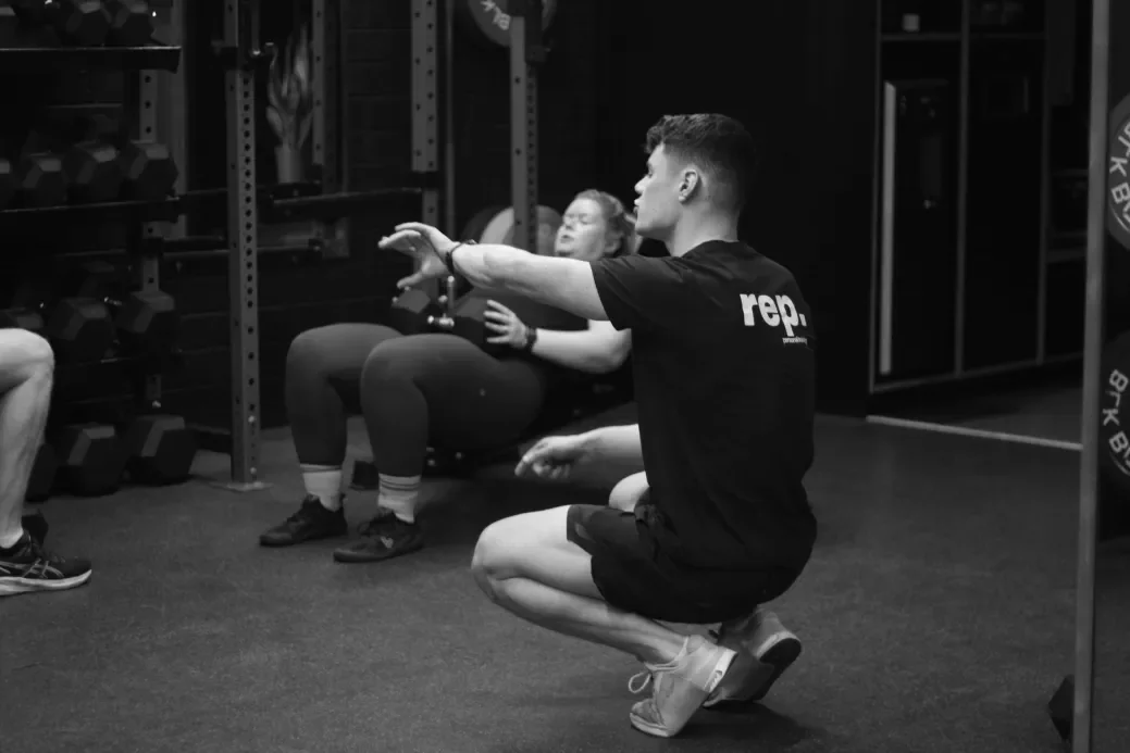 A man squatting and demonstrating an exercise in a gym with two women sitting behind him, with weights and gym equipment in the background.