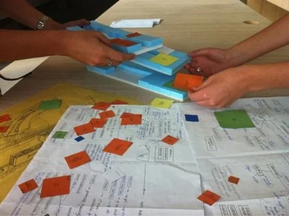 People working on a tabletop with colorful cardboard pieces, papers, and diagrams, assembling or planning a project.