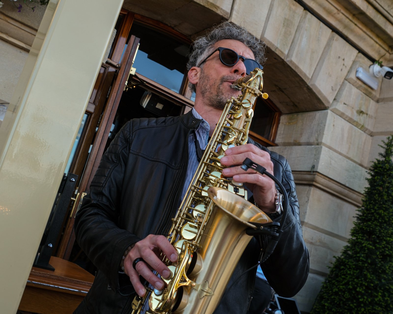A man wearing sunglasses and a leather jacket playing a gold-colored saxophone outdoors in front of a building with stone walls.