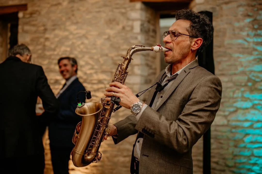 Man playing saxophone at an indoor event with brick wall background and two other men in the background.