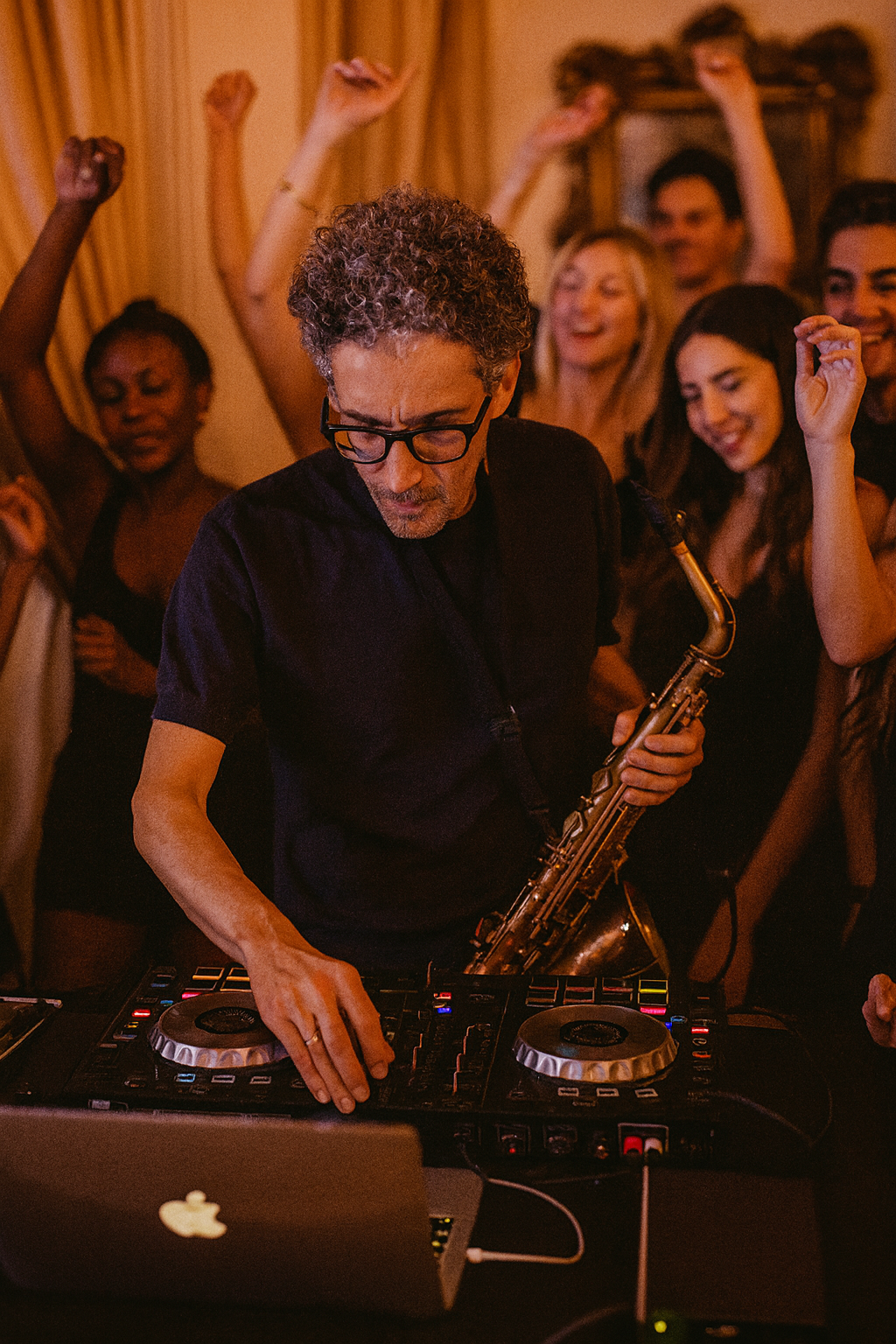 A man playing a DJ controller and a saxophone at a party with several people dancing and celebrating in the background.