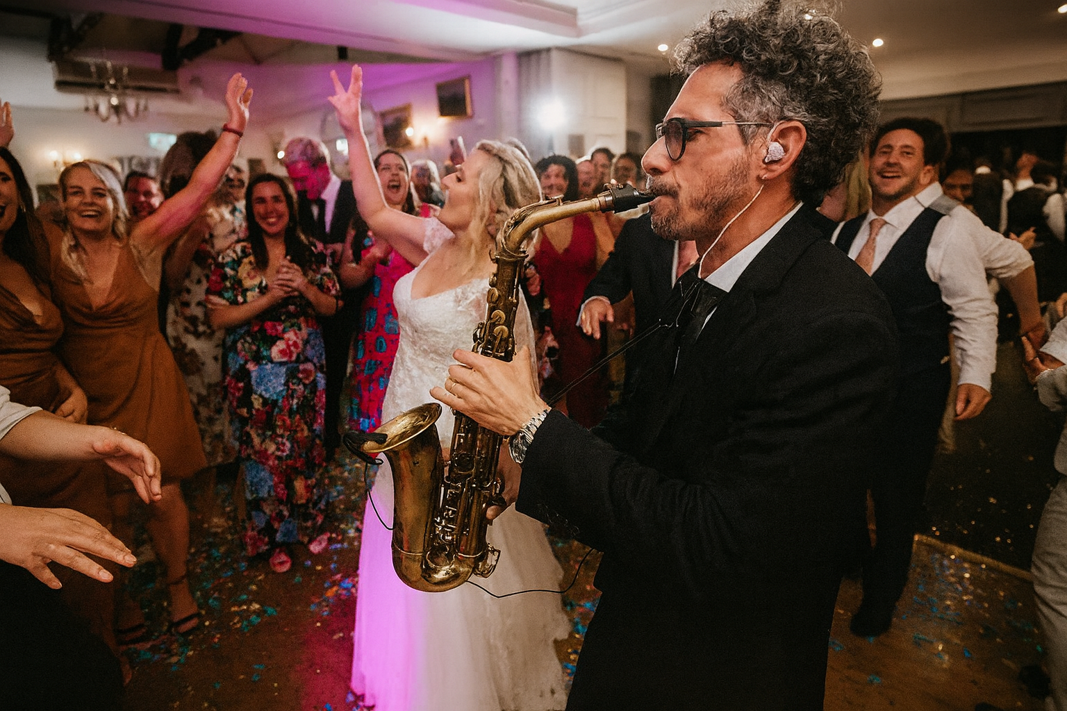 A man playing the saxophone at a lively party or celebration with dancing guests in the background.